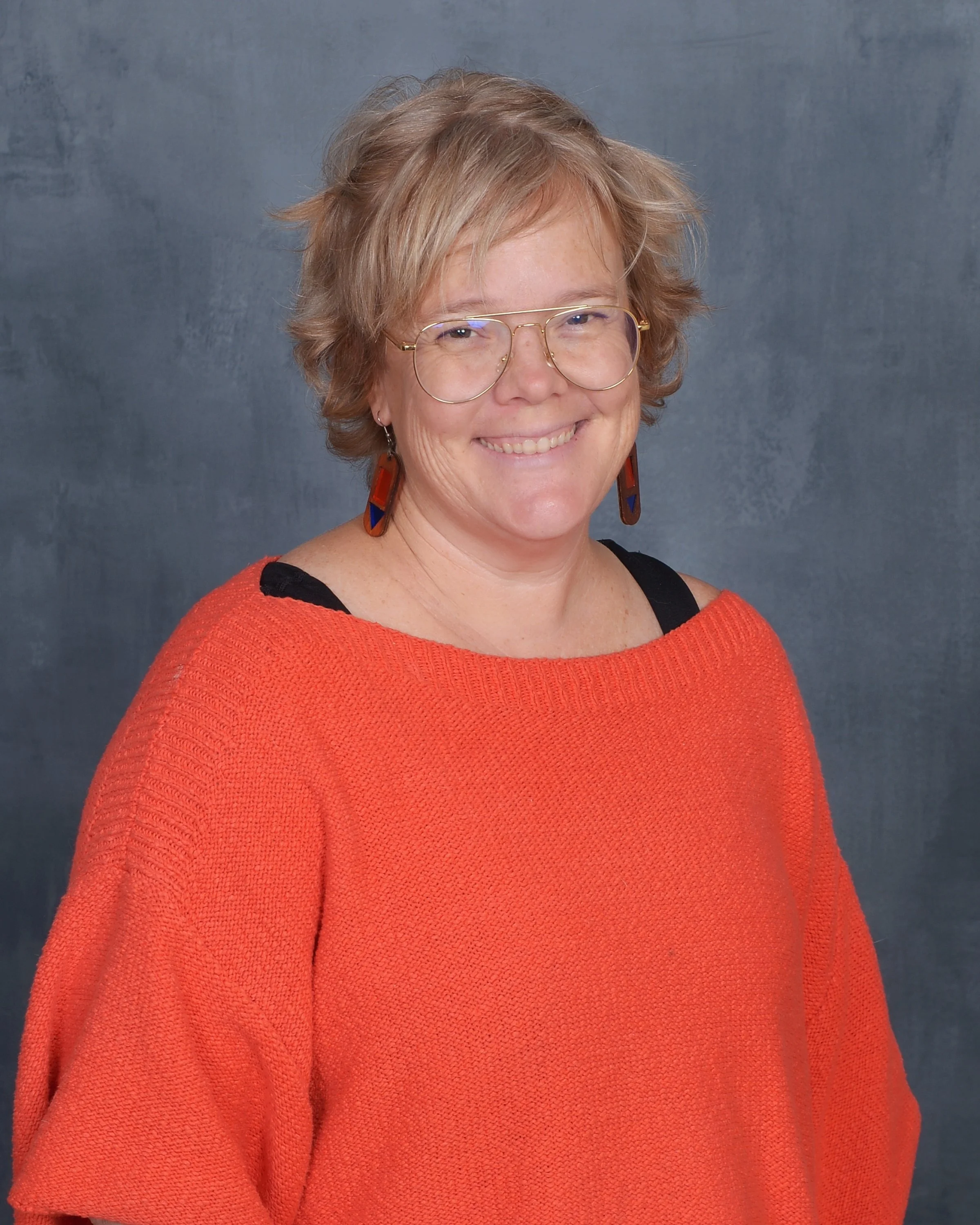 A smiling woman with short, light brown hair and glasses, wearing a red sweater with black accents, standing against a textured gray background.