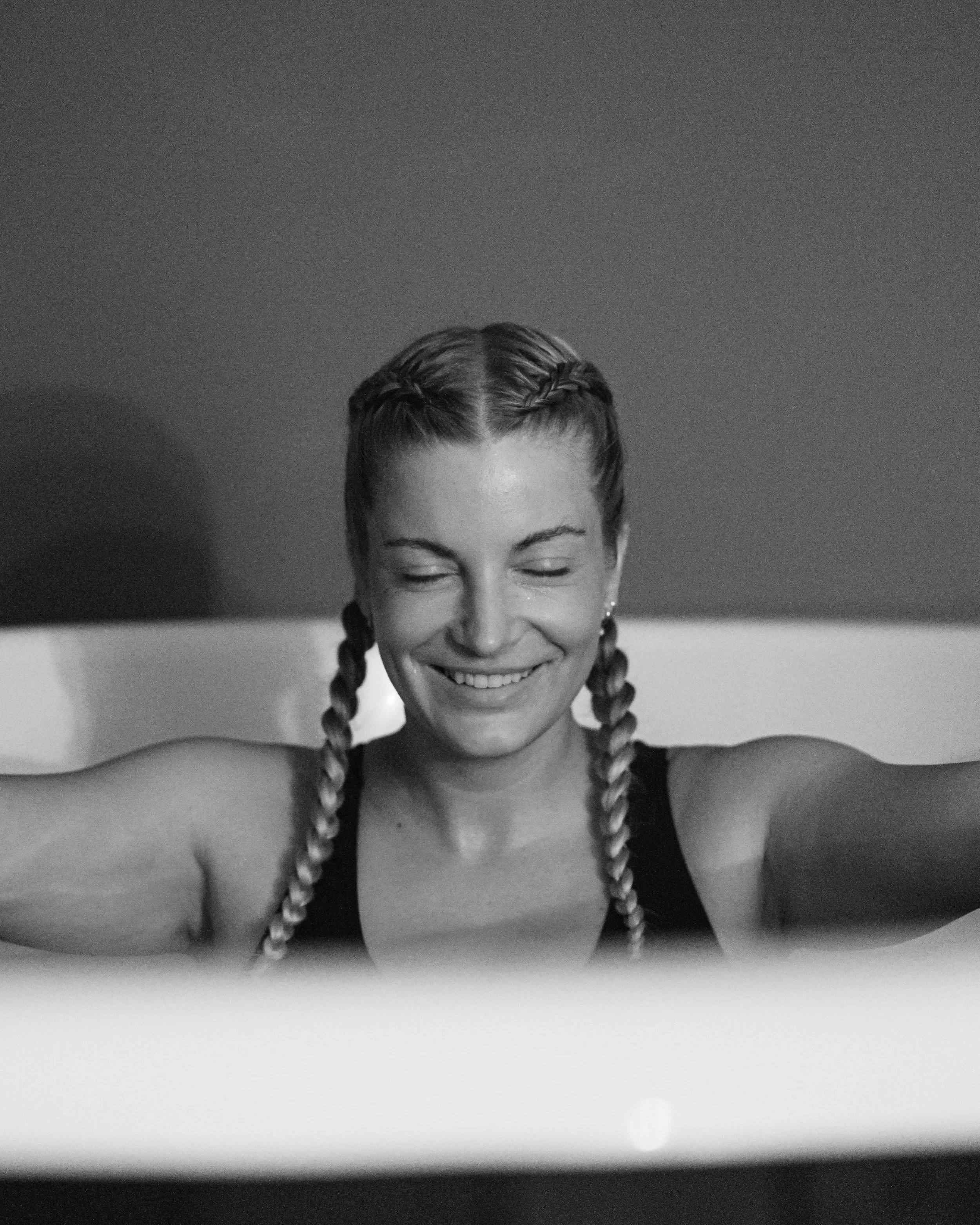 A young woman with braided hair smiling with eyes closed, taking a selfie in black and white.