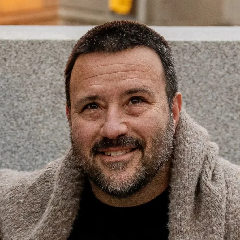 A smiling man with short dark hair and a beard, wearing a cozy gray jacket, sitting outdoors near a gray textured wall.