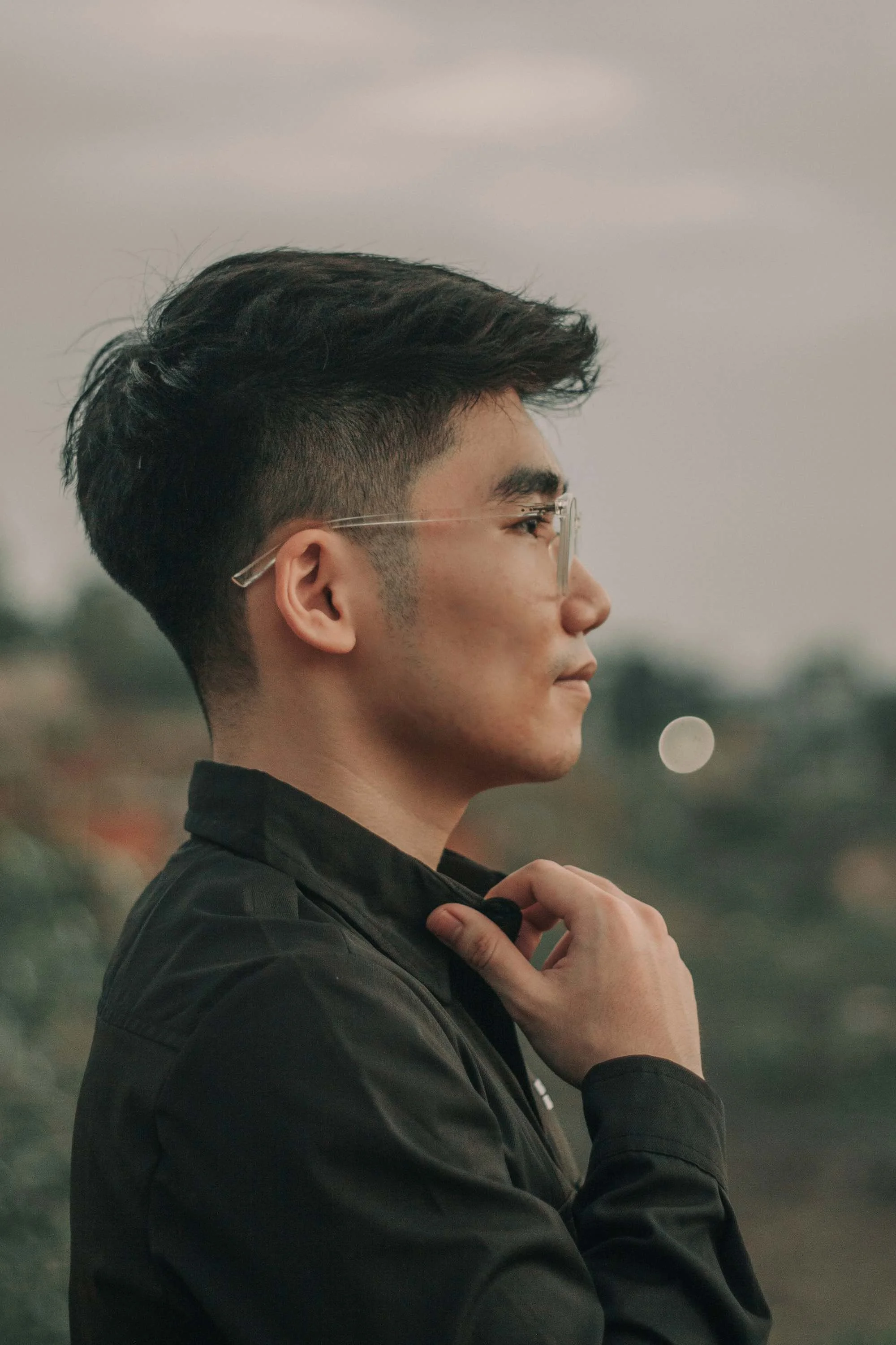 Profile shot of a young man with dark hair, wearing glasses and a black jacket, outdoors with a blurred background.