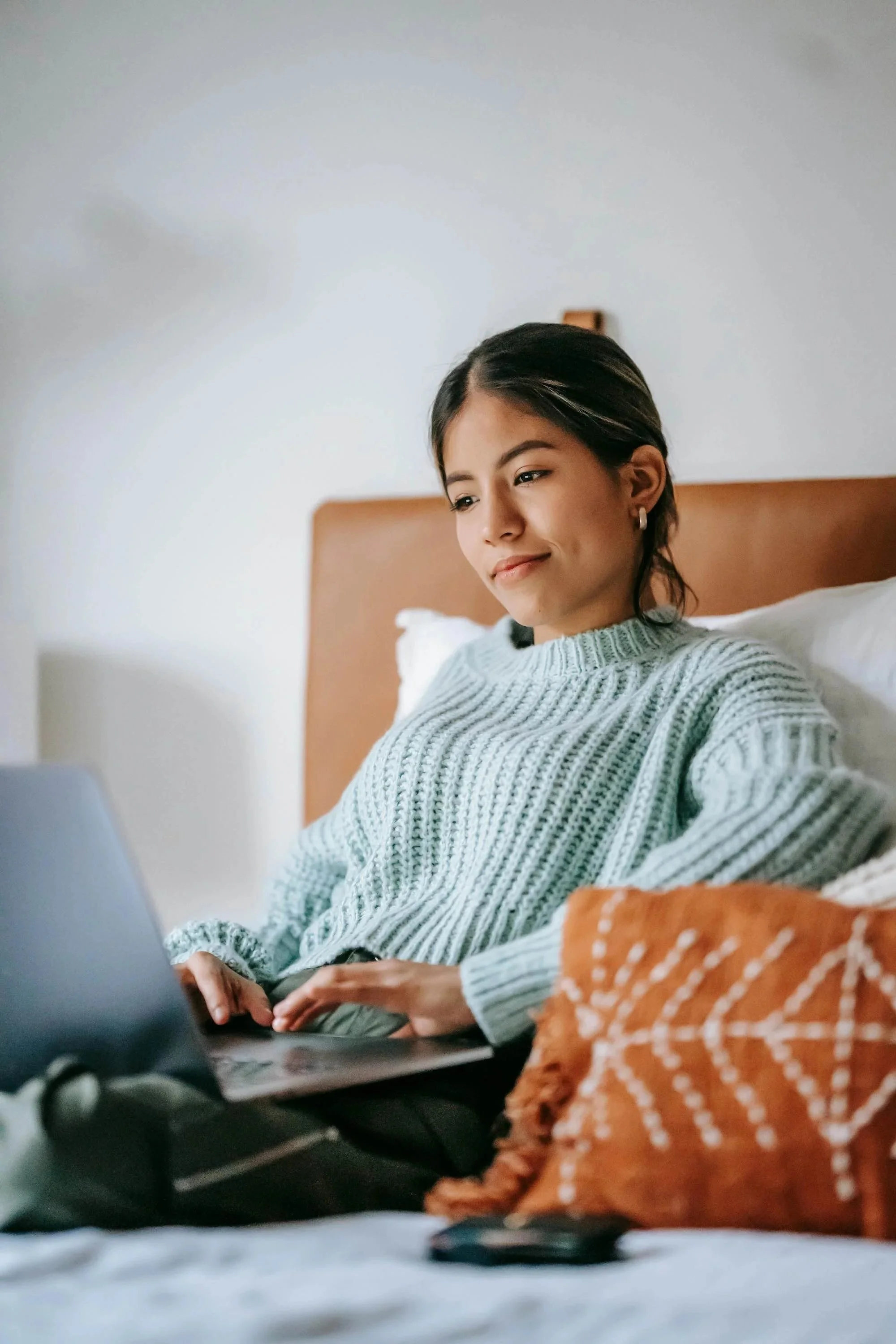 A young woman sitting in bed using a laptop, with a pillow and orange blanket nearby.