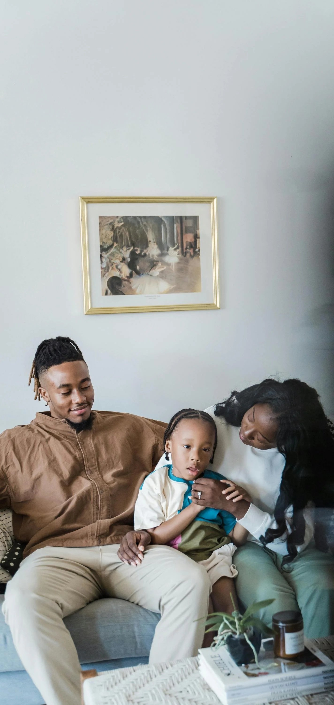 Family sitting on a gray couch, mother holding young boy, father sitting beside them, in a living room with art painting on white wall, coffee table with books, plant, and candle.