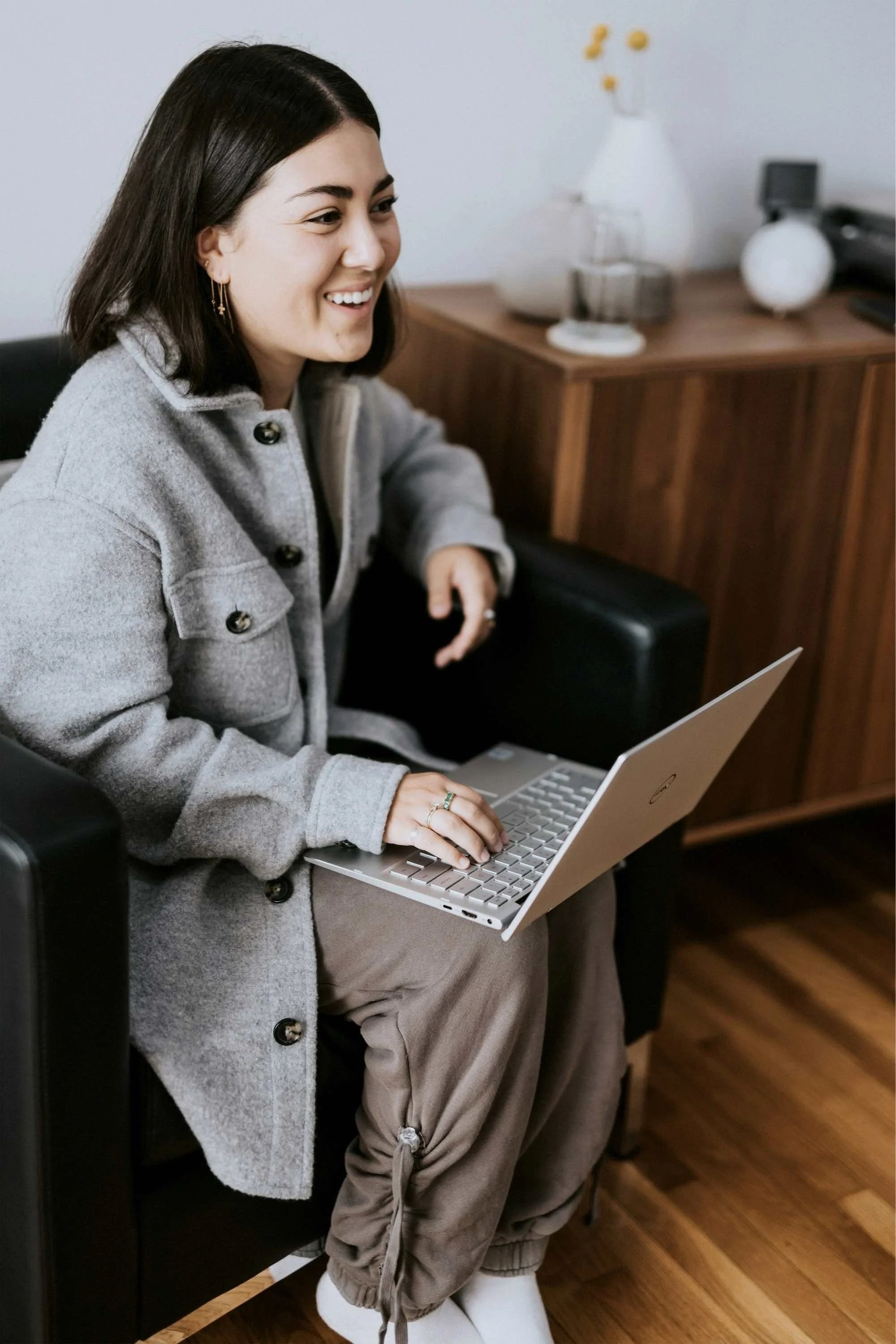 Woman sitting on a black chair, smiling and using a silver laptop in a cozy room with wooden furniture and decorative vases.