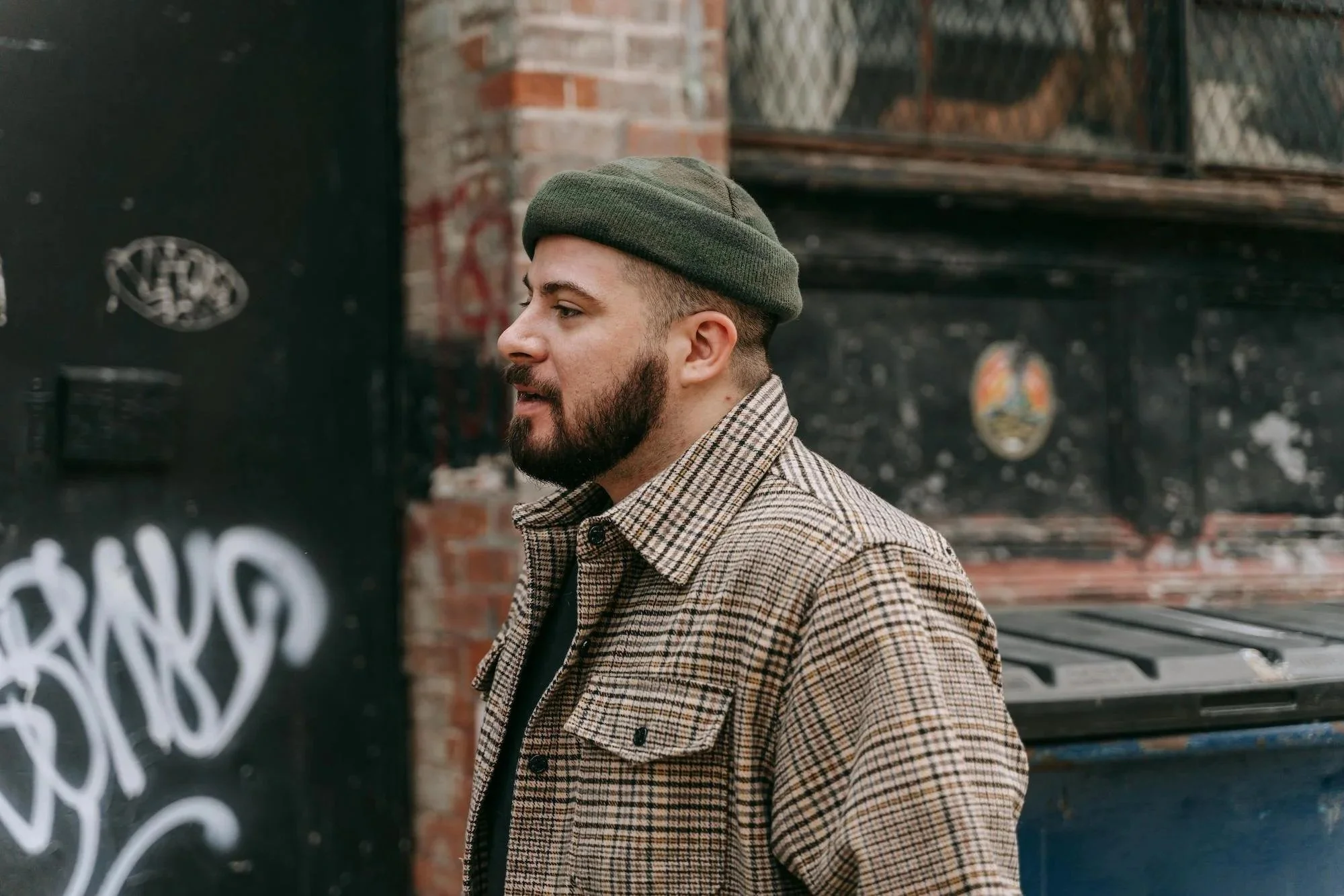 A man with a beard wearing a green beanie and a checked jacket stands next to a brick wall with graffiti.