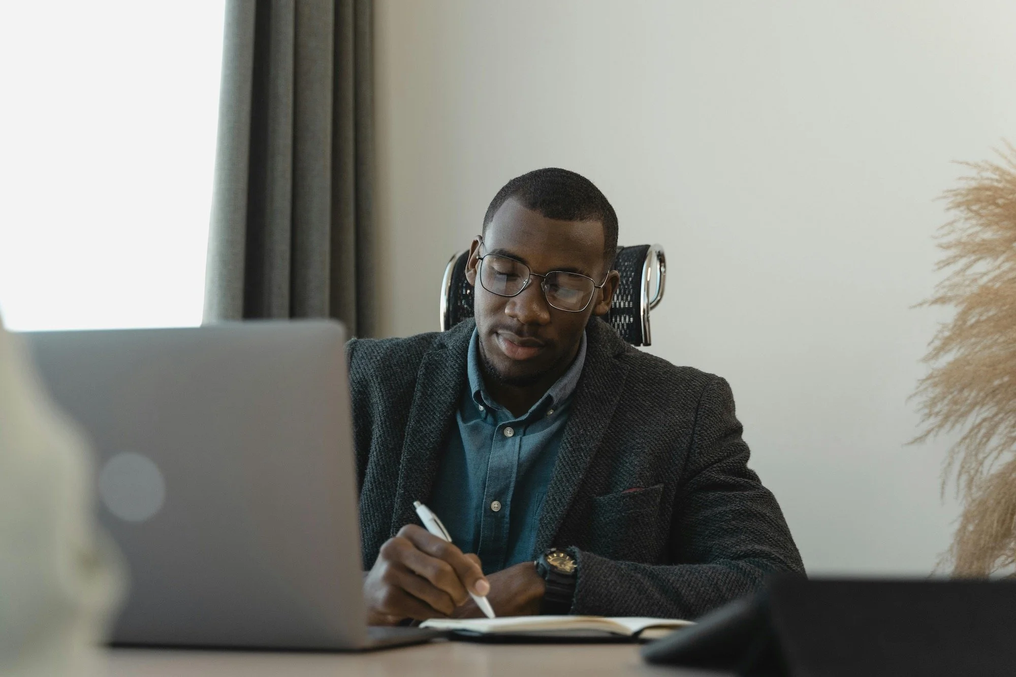 A man with glasses and a dark blazer working at his desk with a laptop and a notebook in an office with curtains and a white wall.