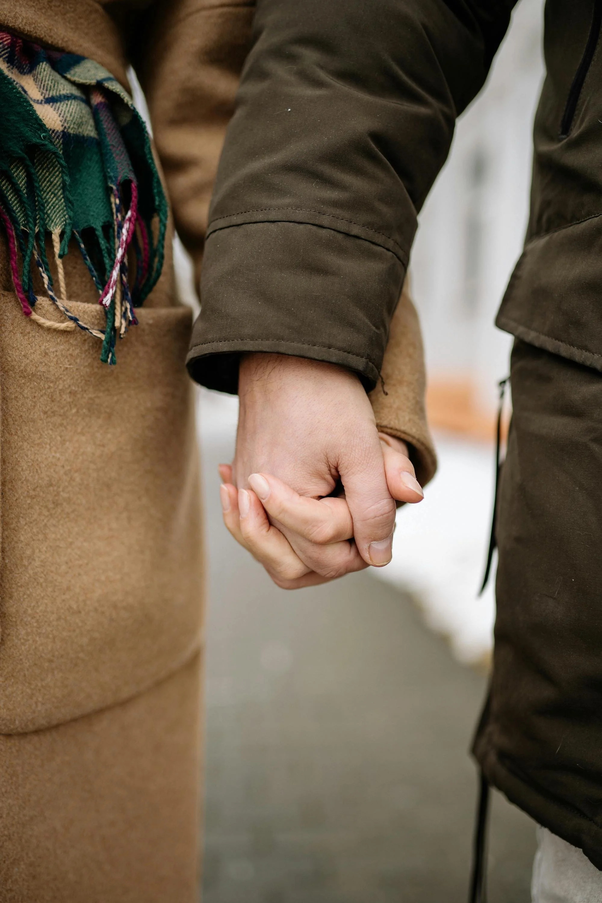 Close-up of two people holding hands, one person wearing a brown coat and a scarf, the other wearing a dark jacket.
