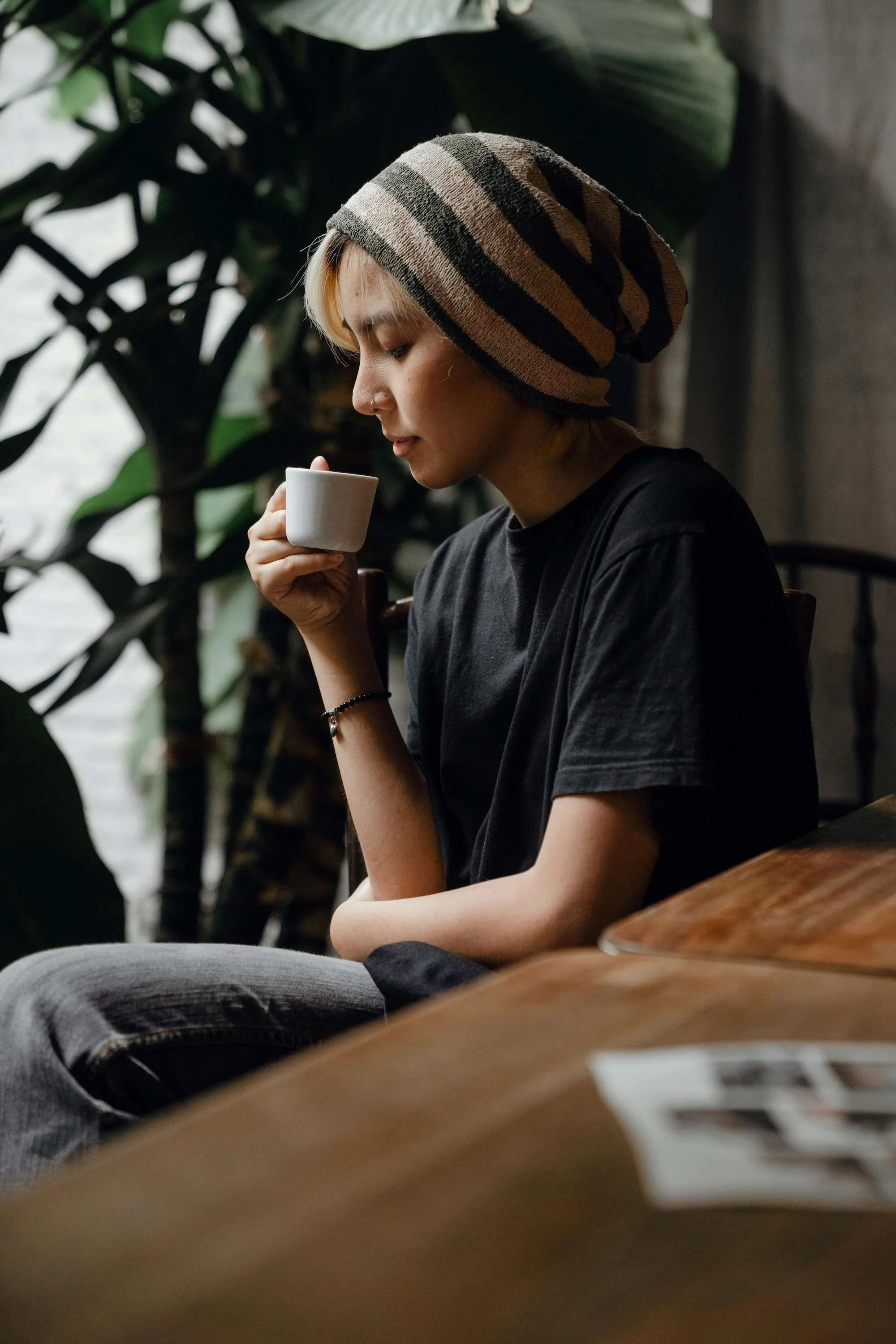 A young person with a striped beanie and nose piercing sitting by a window, holding a white mug, in a cozy, leafy cafe.