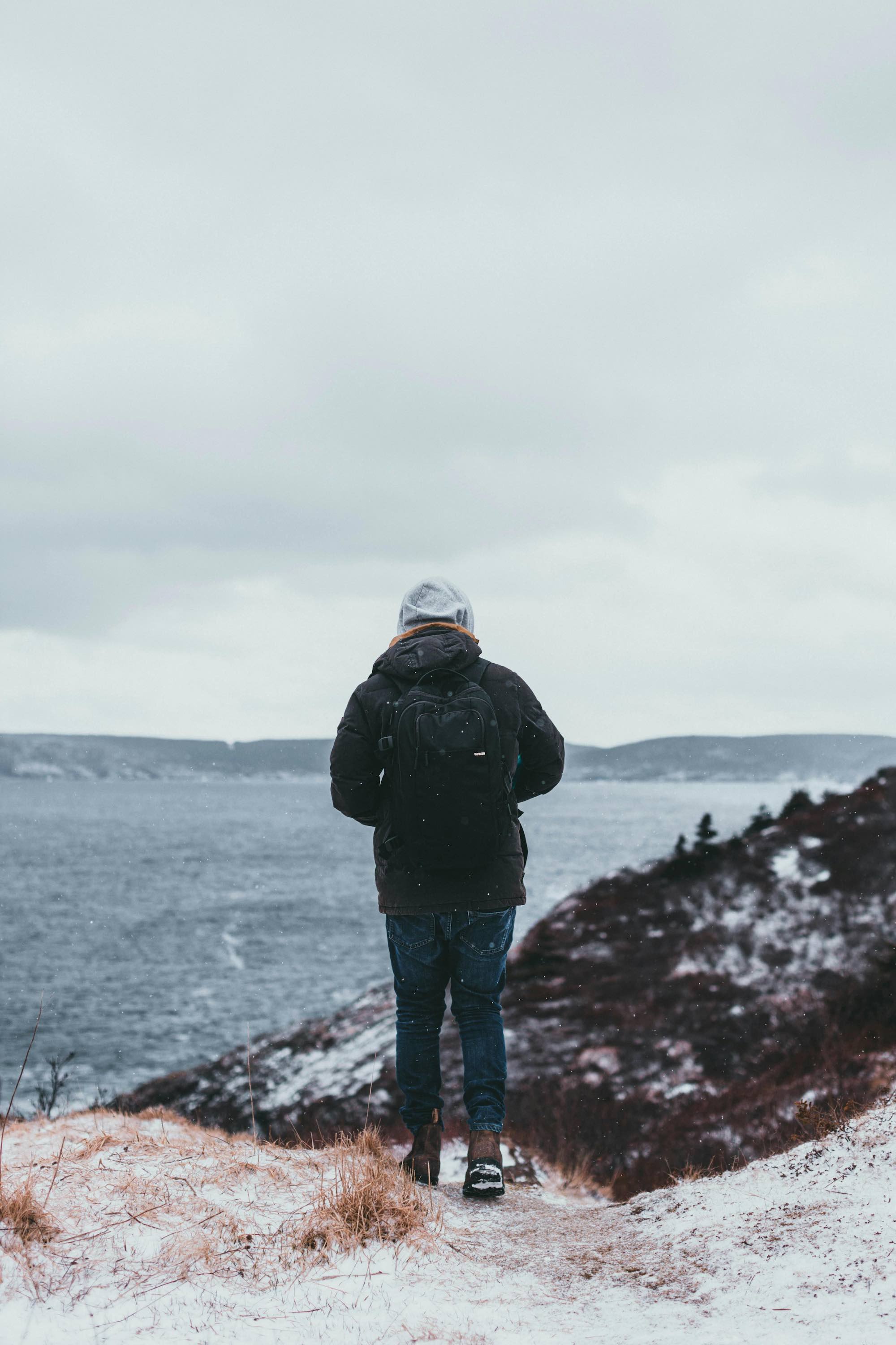 Person in a black jacket, jeans, and gray beanie hiking near a lake in a snowy landscape.