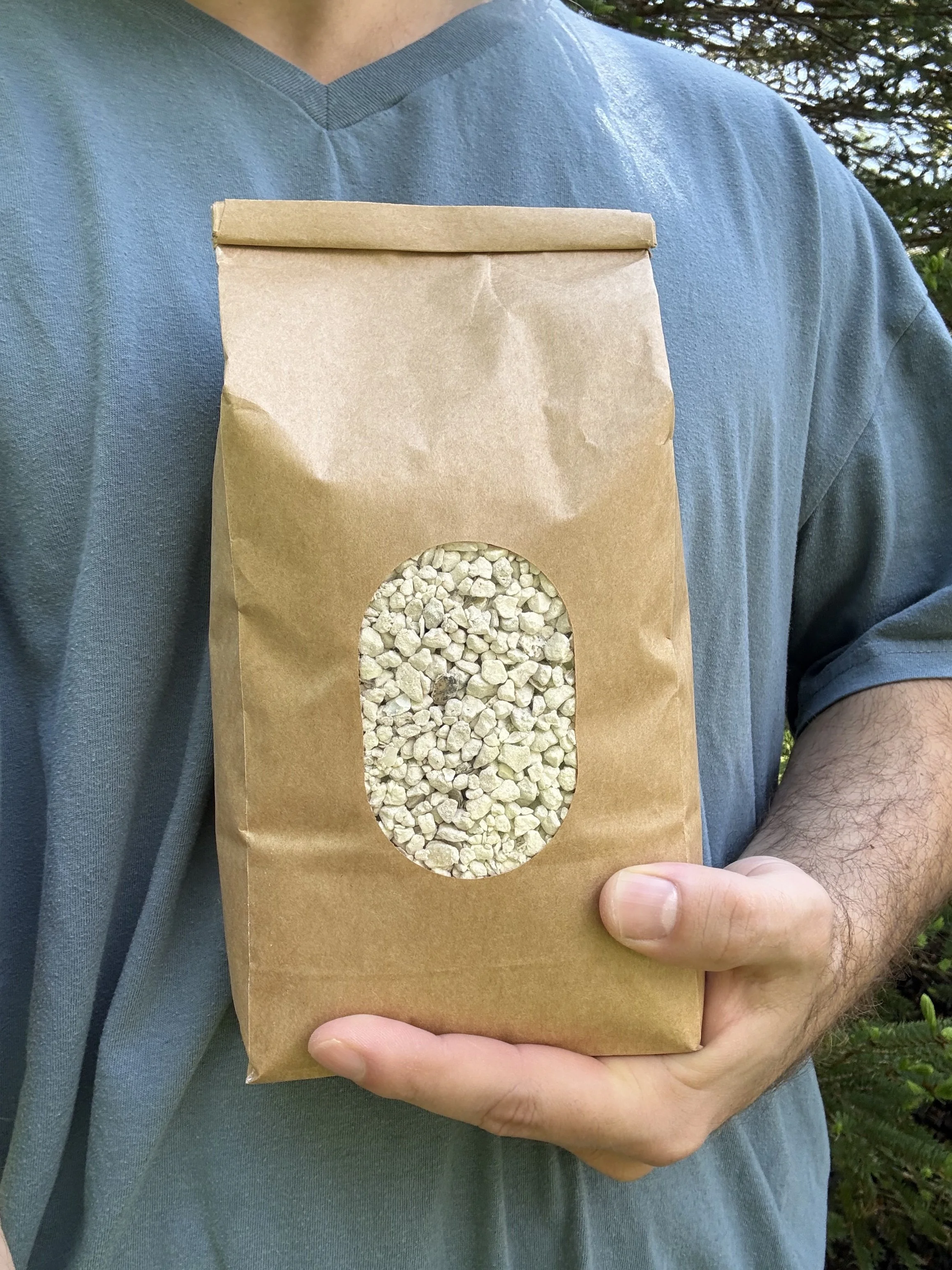 A person wearing a blue shirt holds a brown paper bag with a clear oval window showing Oxwhale's crushed oyster shells inside.