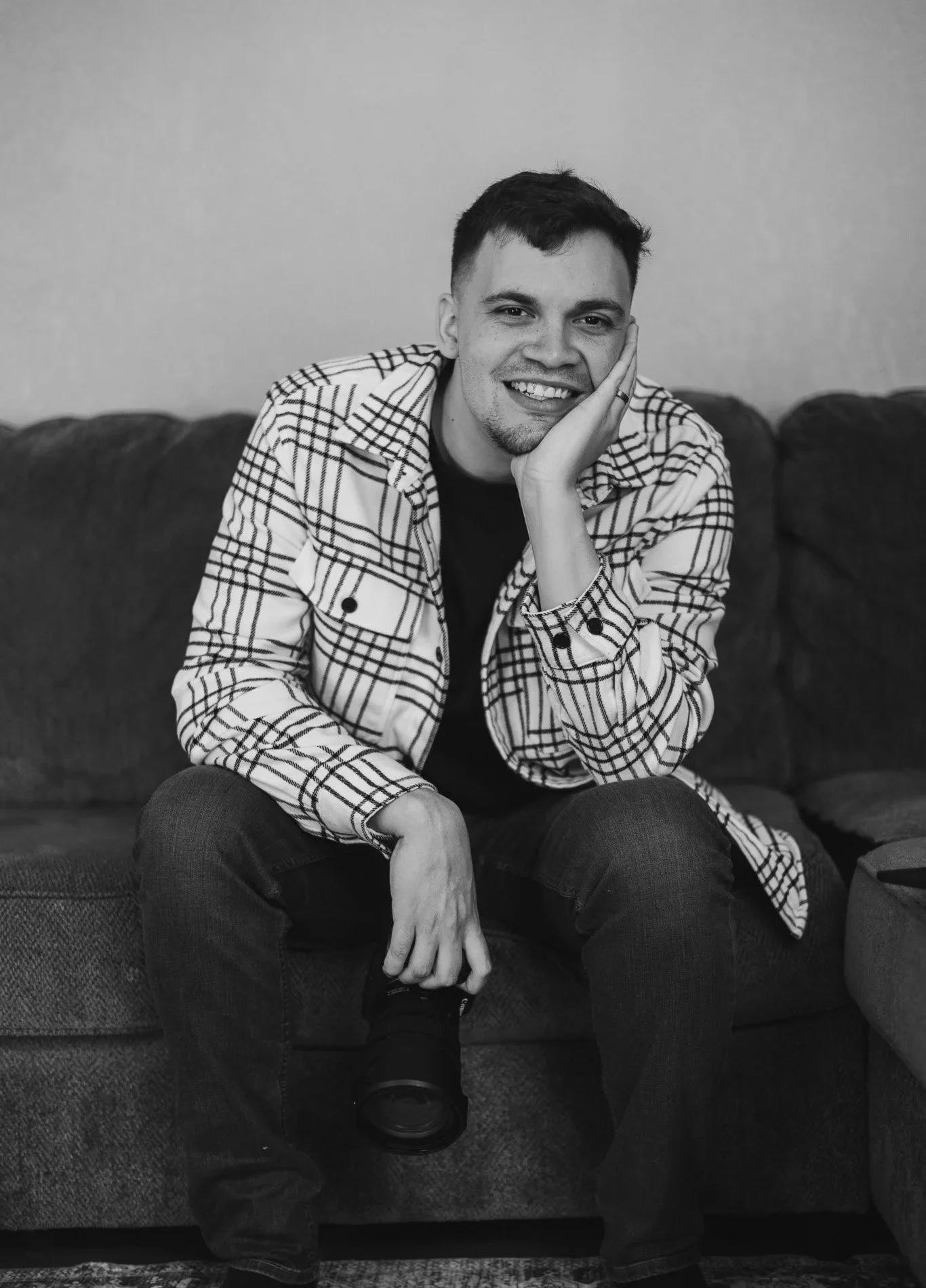 A smiling young man sitting on a sofa, resting his face on one hand, dressed in a checkered jacket and dark jeans, with a camera in his other hand.