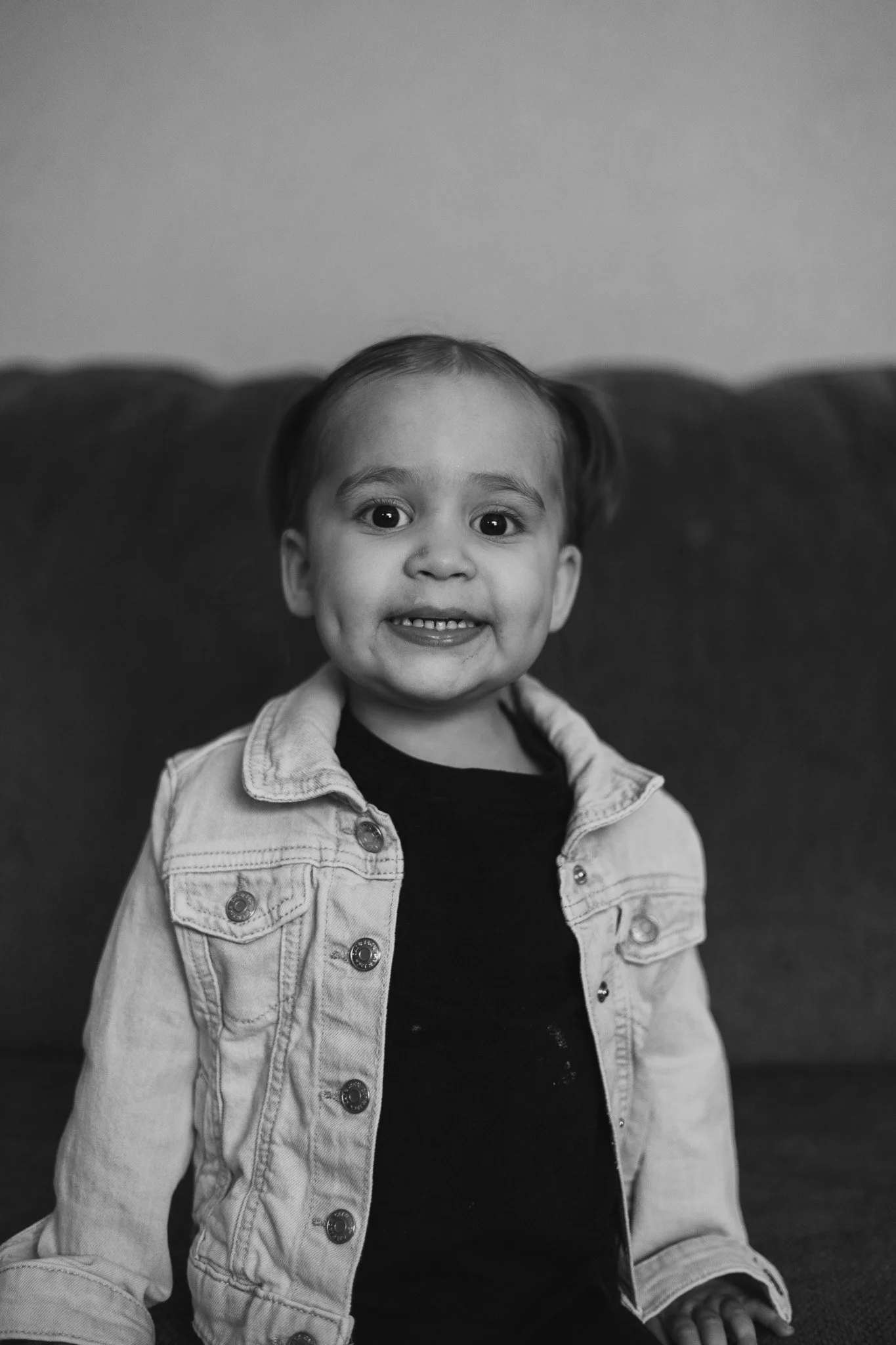 Young girl with a big smile, wearing a denim jacket and a black shirt, sitting in front of a plain background.