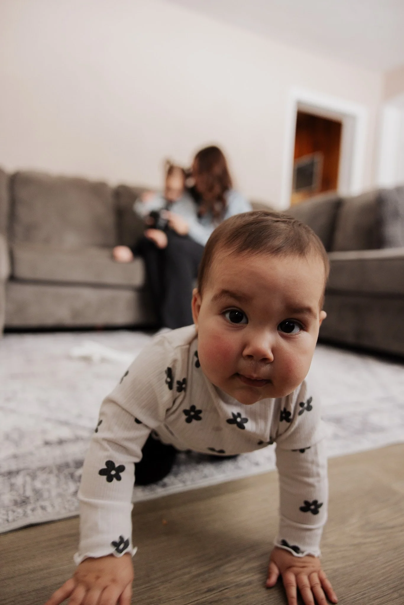 A young child with short brown hair crawling on a wooden floor, wearing a beige sweater with black flowers. In the background, a woman sitting on a gray couch holding a baby.