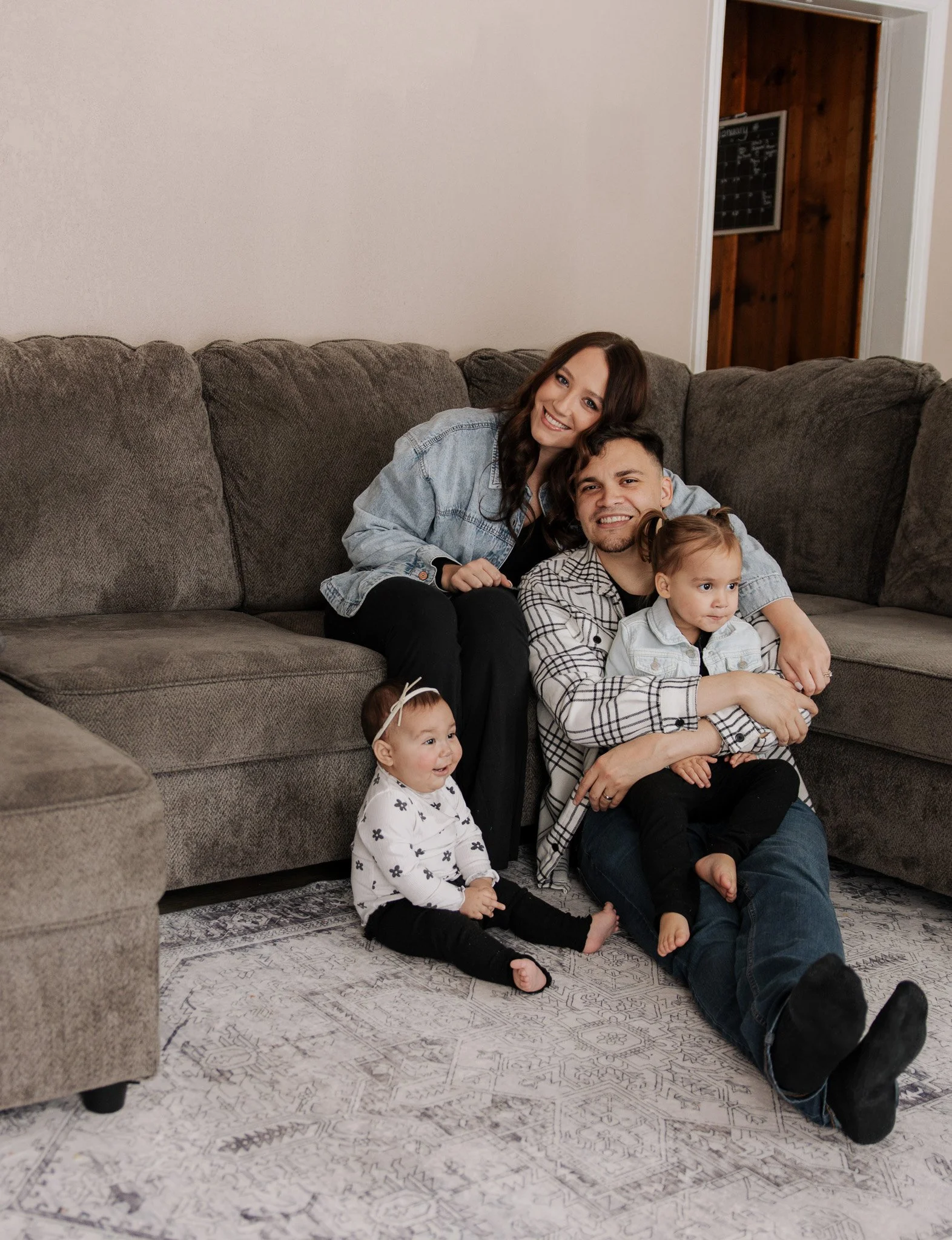 Family of four sitting together on living room floor and couch, smiling and enjoying quality time.