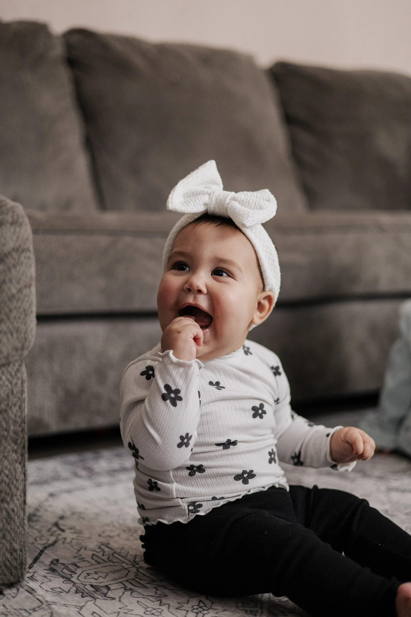 A baby girl with a white headband with a bow, sitting on the floor, wearing a white shirt with black flowers and black pants, in a living room with a gray sofa in the background.
