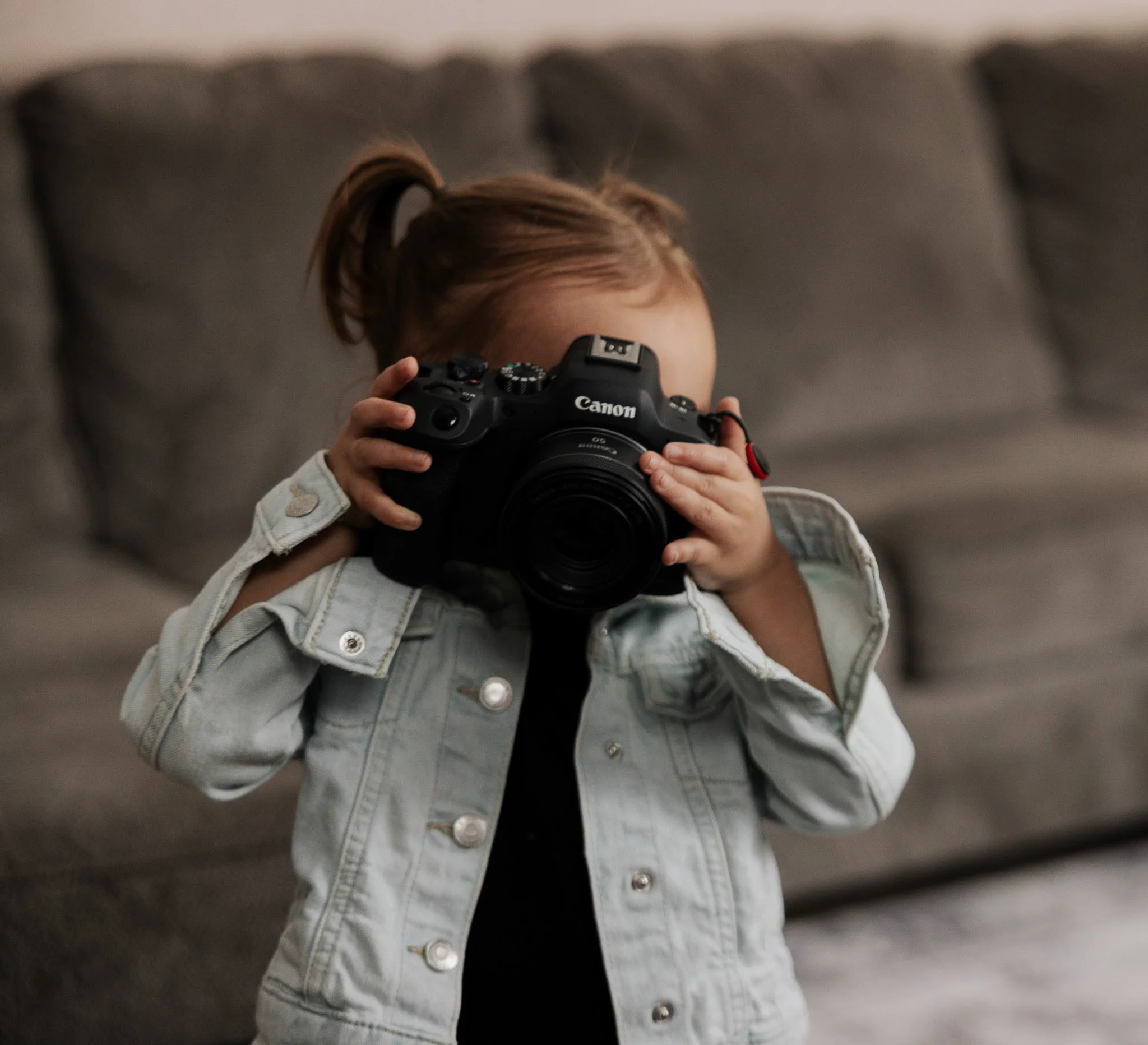 A young child with light brown hair tied in a ponytail holds a Canon camera up to her face, standing in front of a grey couch.