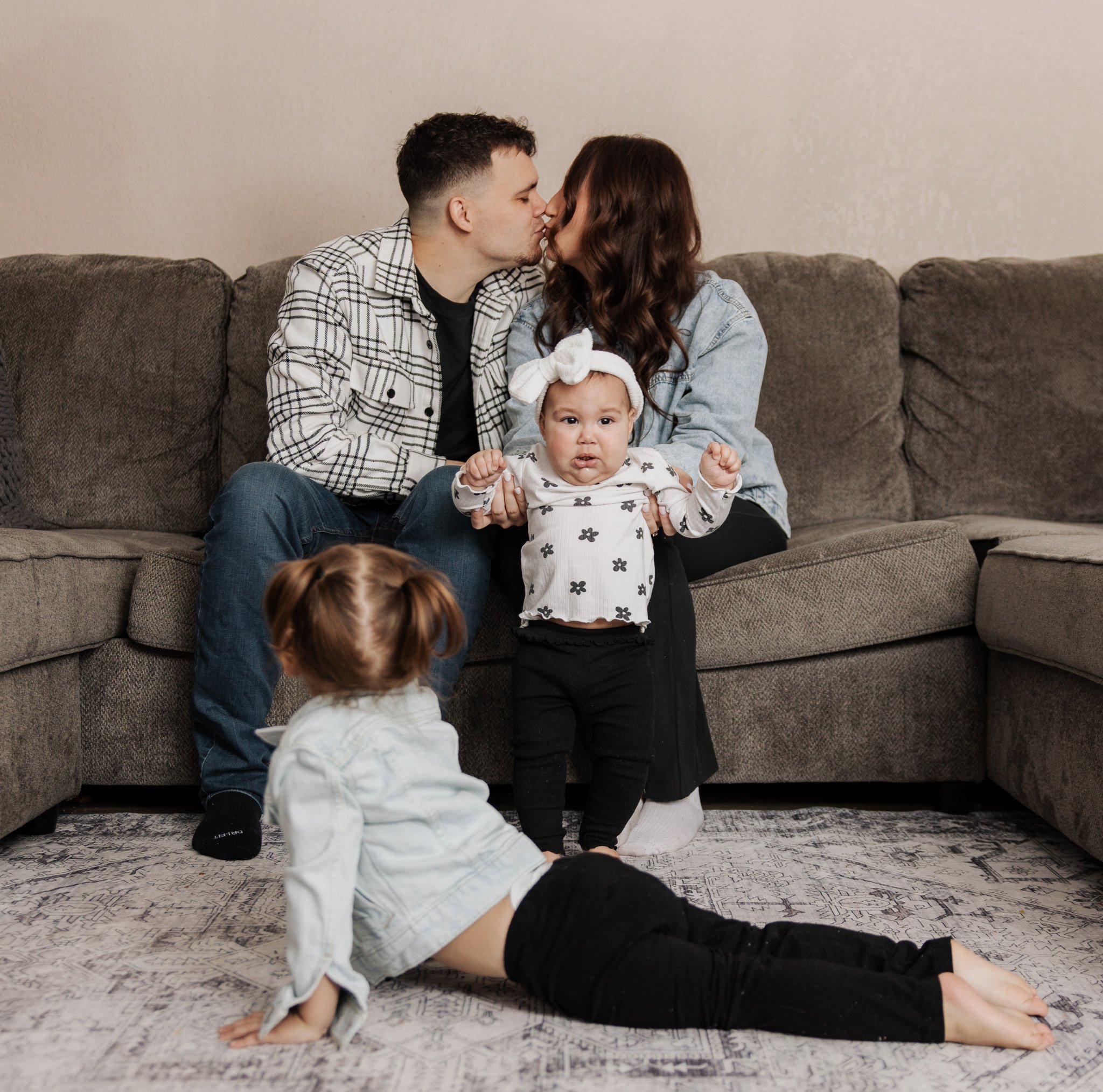 A couple sitting on a couch kissing, with two young children nearby. One child is standing, holding the parent's hand, and the other is lying on the floor.
