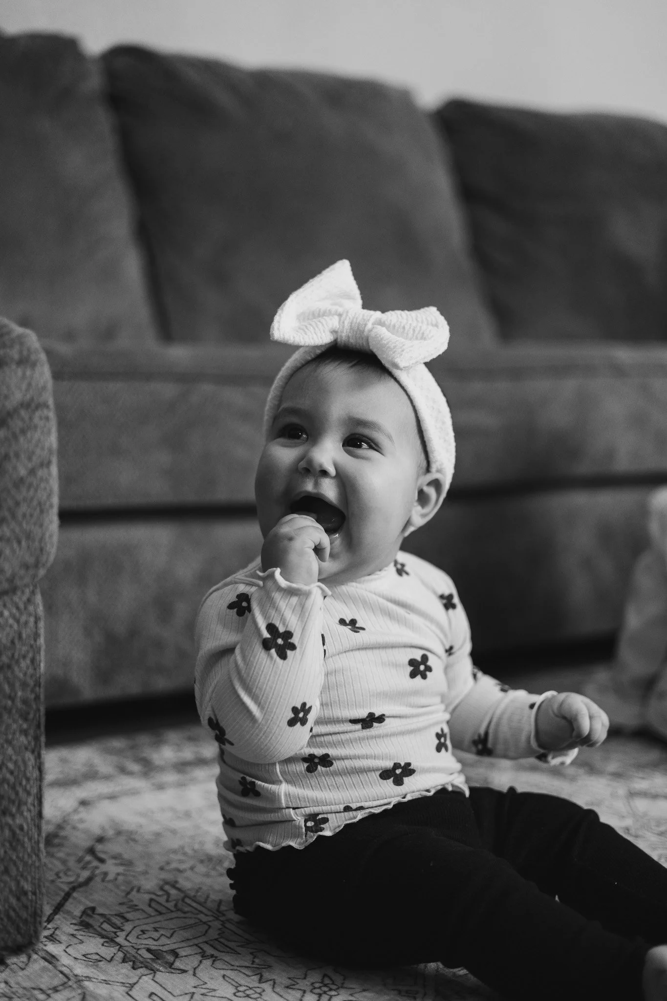 A young girl with a headband and a bow, sitting on a carpet in a living room, smiling and touching her face.