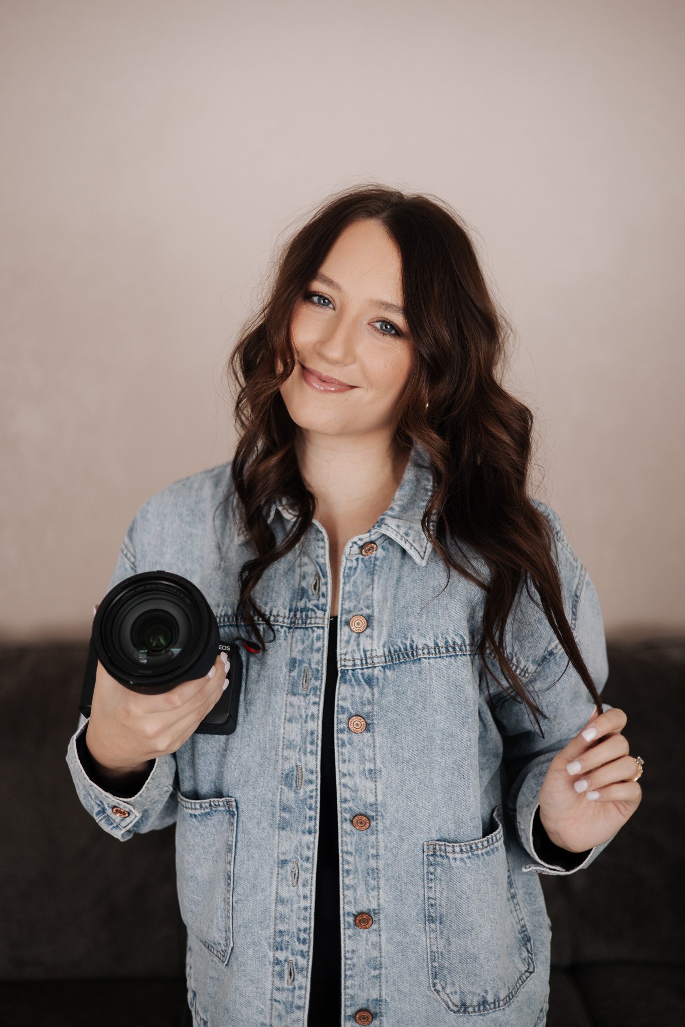 A young woman with long brown wavy hair wearing a denim jacket, holding a camera, smiling, indoors with a beige background.