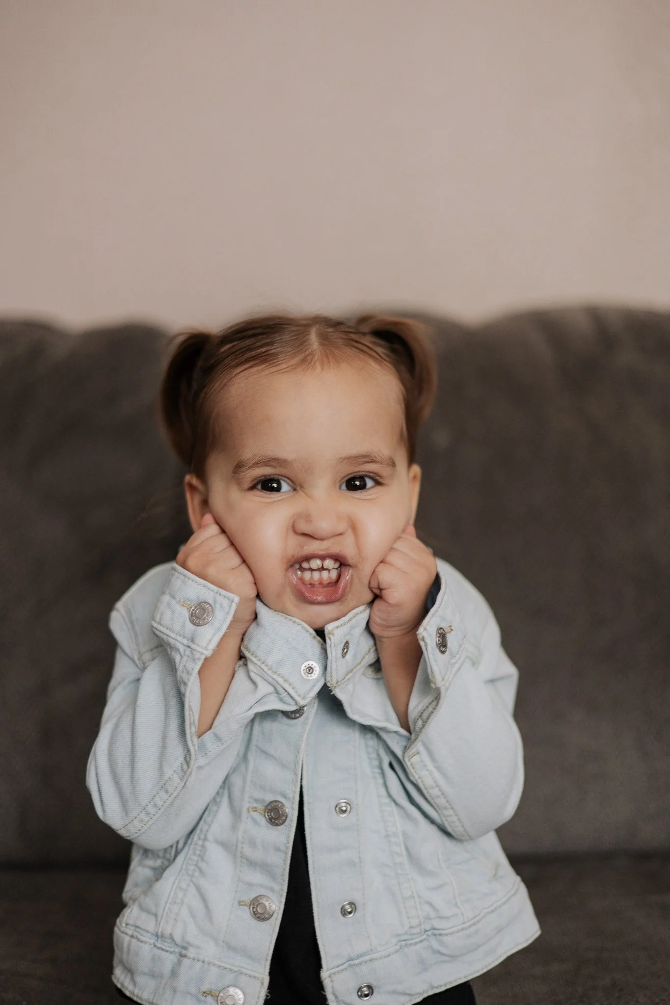 A young girl with light brown hair in pigtails, making a funny face with clenched fists, showing her teeth and scrunching her nose, wearing a light denim jacket, sitting on a dark gray couch.