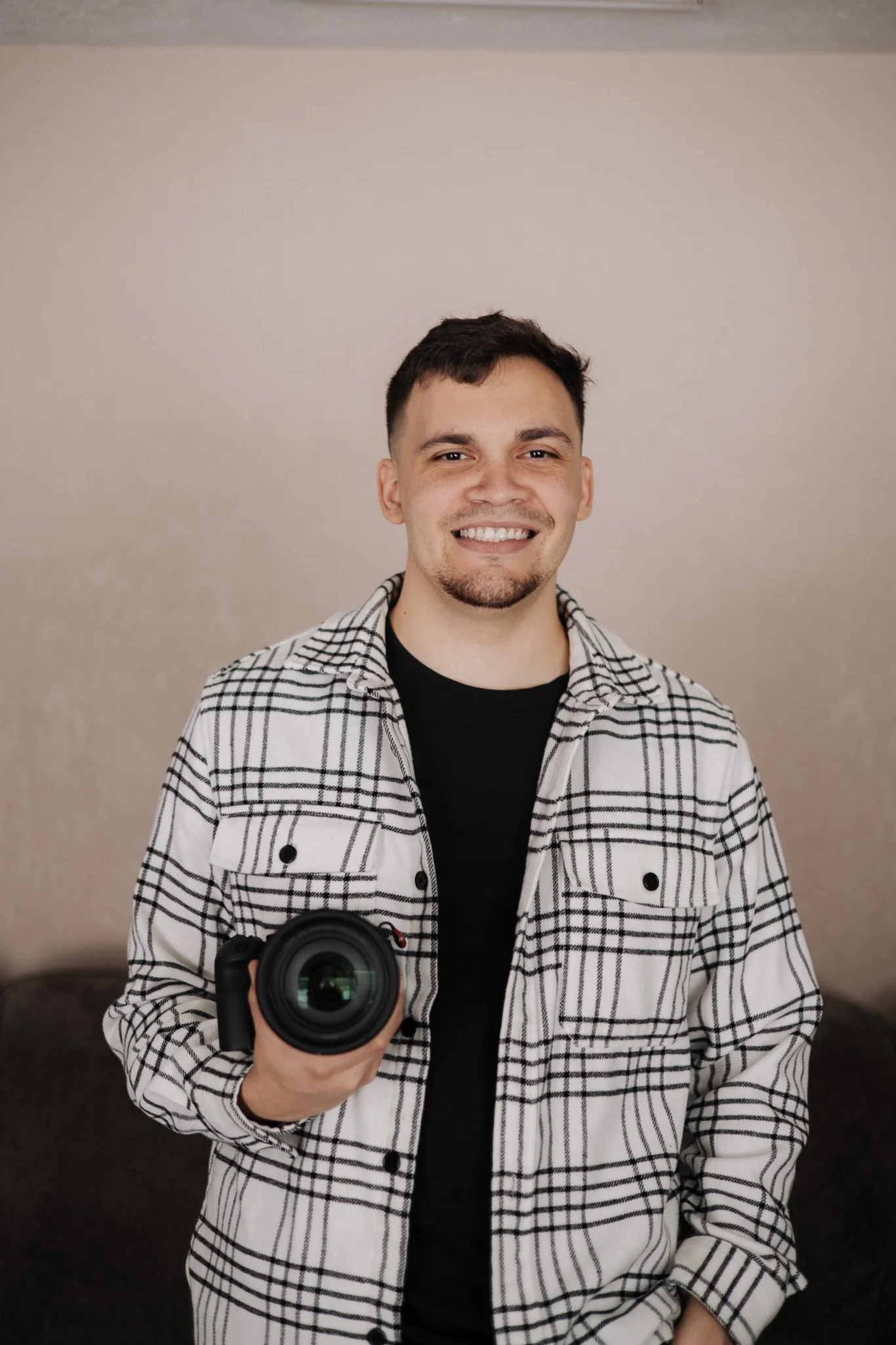 A young man with short dark hair, a light beard, and a big smile, holding a camera in his right hand, wearing a white and black plaid shirt over a black t-shirt, standing against a plain beige wall.