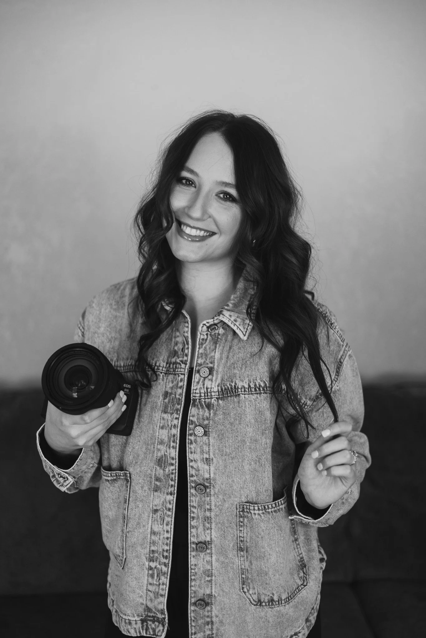 A woman with long wavy hair smiling, holding a camera in her right hand, wearing a denim jacket.