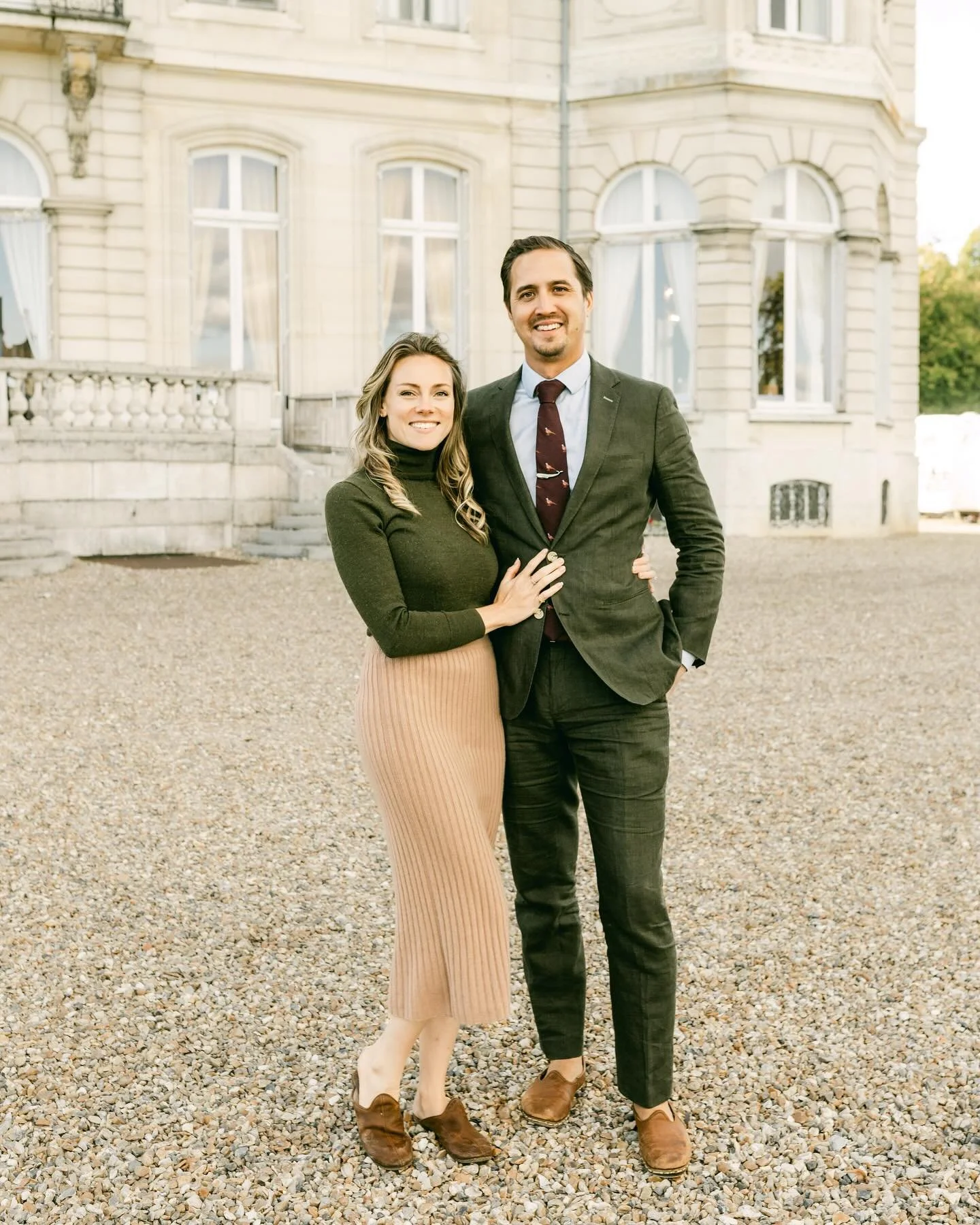 A man and woman standing outside in front of an elegant, historic building with large arched windows, dressed in business casual attire, smiling at the camera.