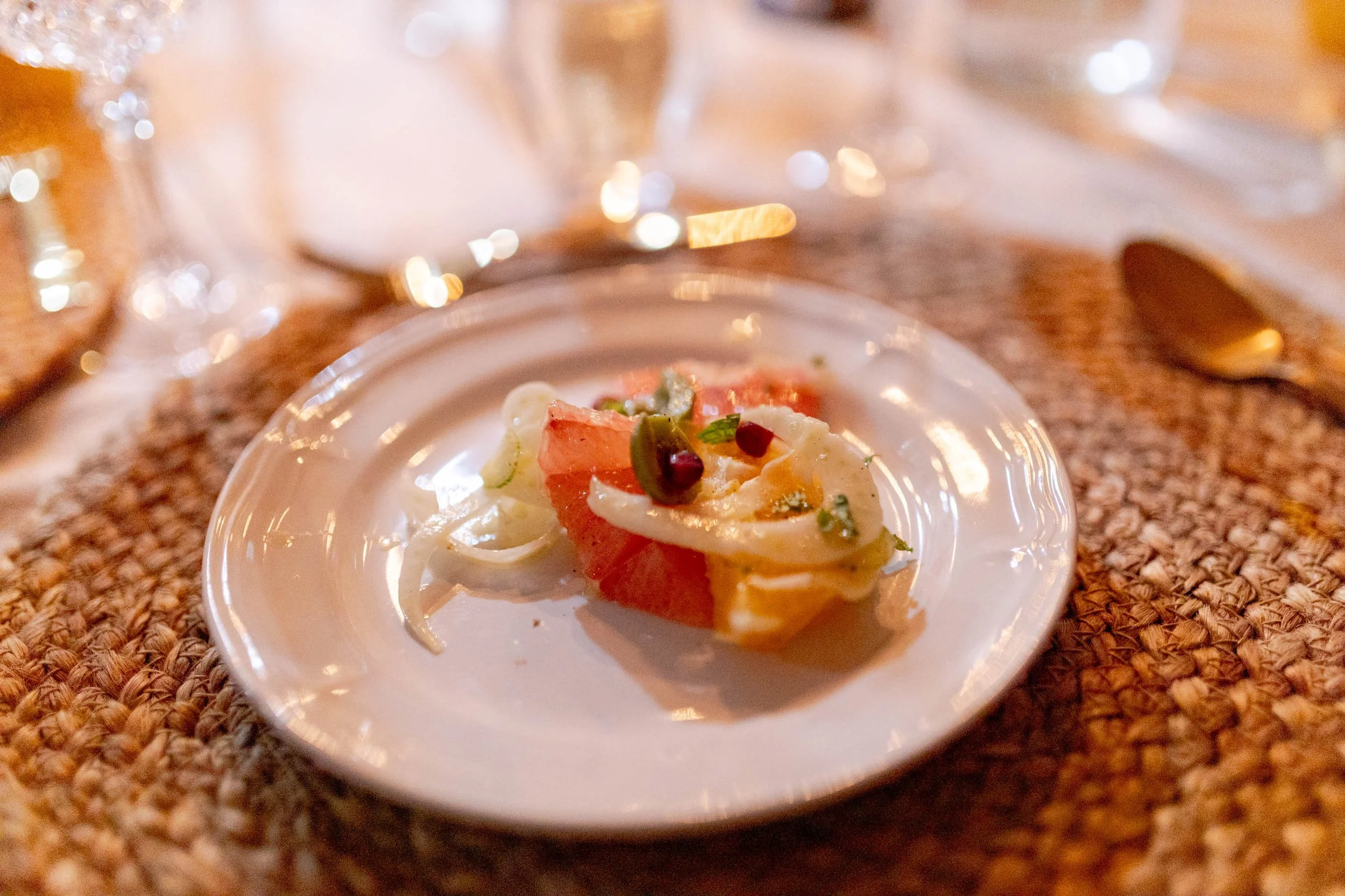 Close-up of a small appetizer on a white plate with sliced onions, watermelon, and herbs, set on a woven placemat with a spoon and glasses in the background.