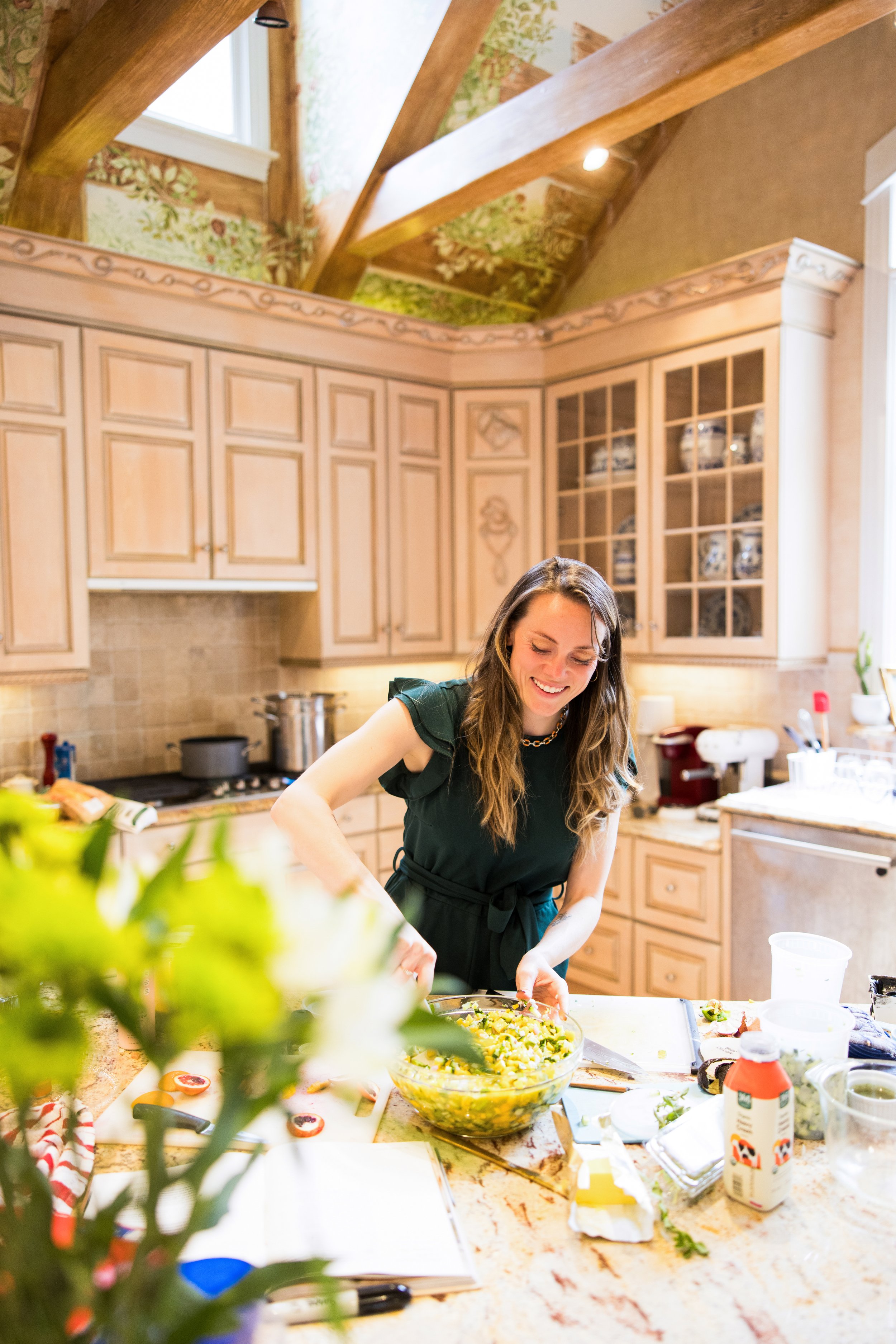 A woman in a black apron smiling and preparing a salad in a bright kitchen with wooden cabinets and a high, sloped ceiling.