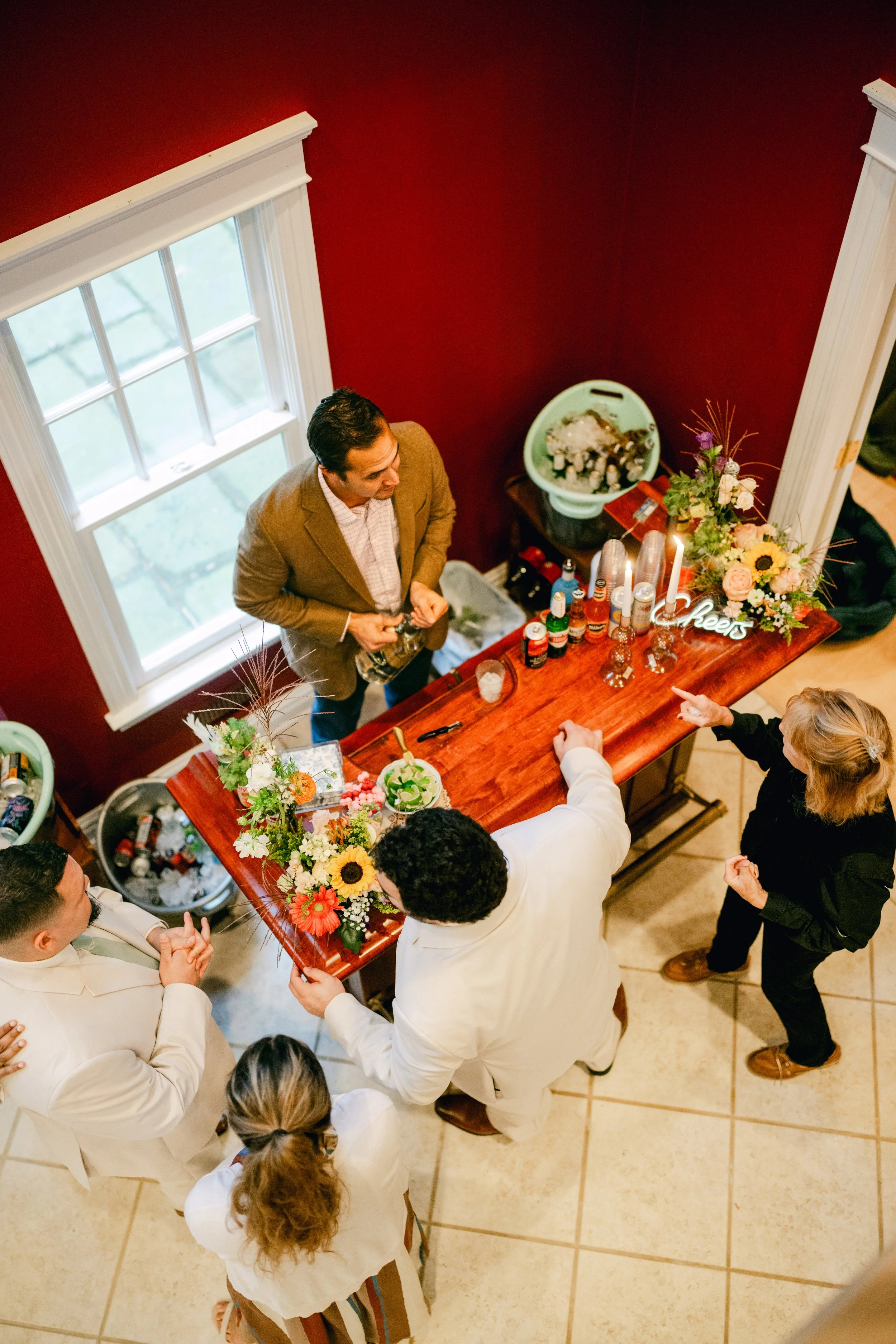 People gathered around a wooden bar table, engaged in conversation, with floral decorations, drinks, and a 