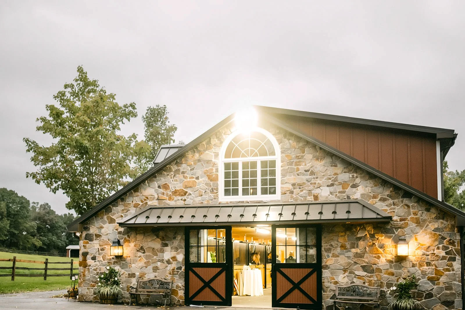 A rustic stone barn with barn doors open, revealing an indoor event space with tables and decorations, surrounded by trees and a partly cloudy sky.