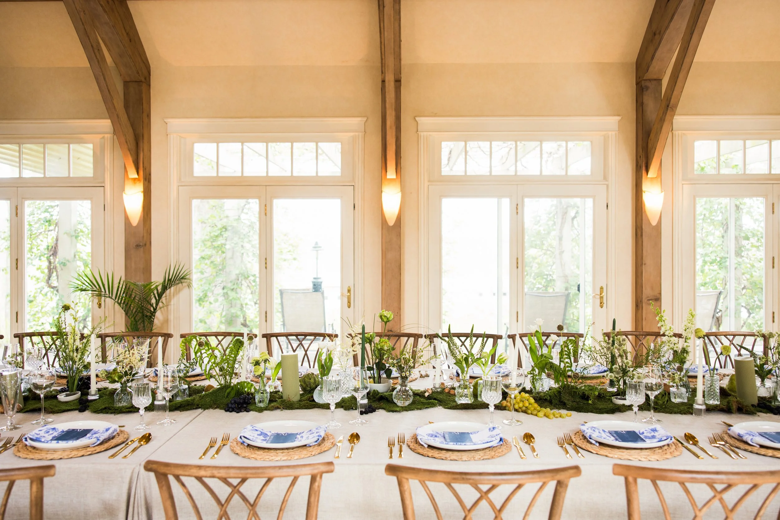Elegant dining table with white tablecloth, gold cutlery, blue and white plates, surrounded by wooden chairs, decorated with green plants and flowers, in a bright room with large windows and wooden beams.