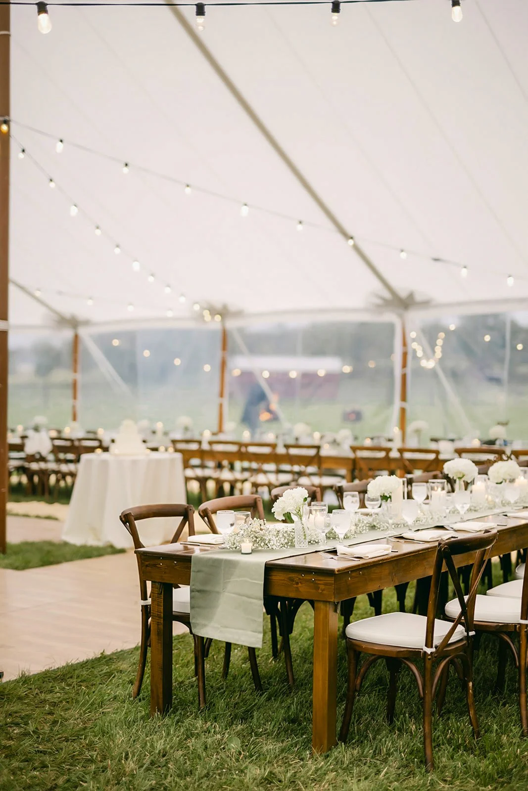 Indoor wedding reception set up with long wooden tables decorated with white flowers and tableware, under a white tent with string lights, on a grassy area with a view outside.