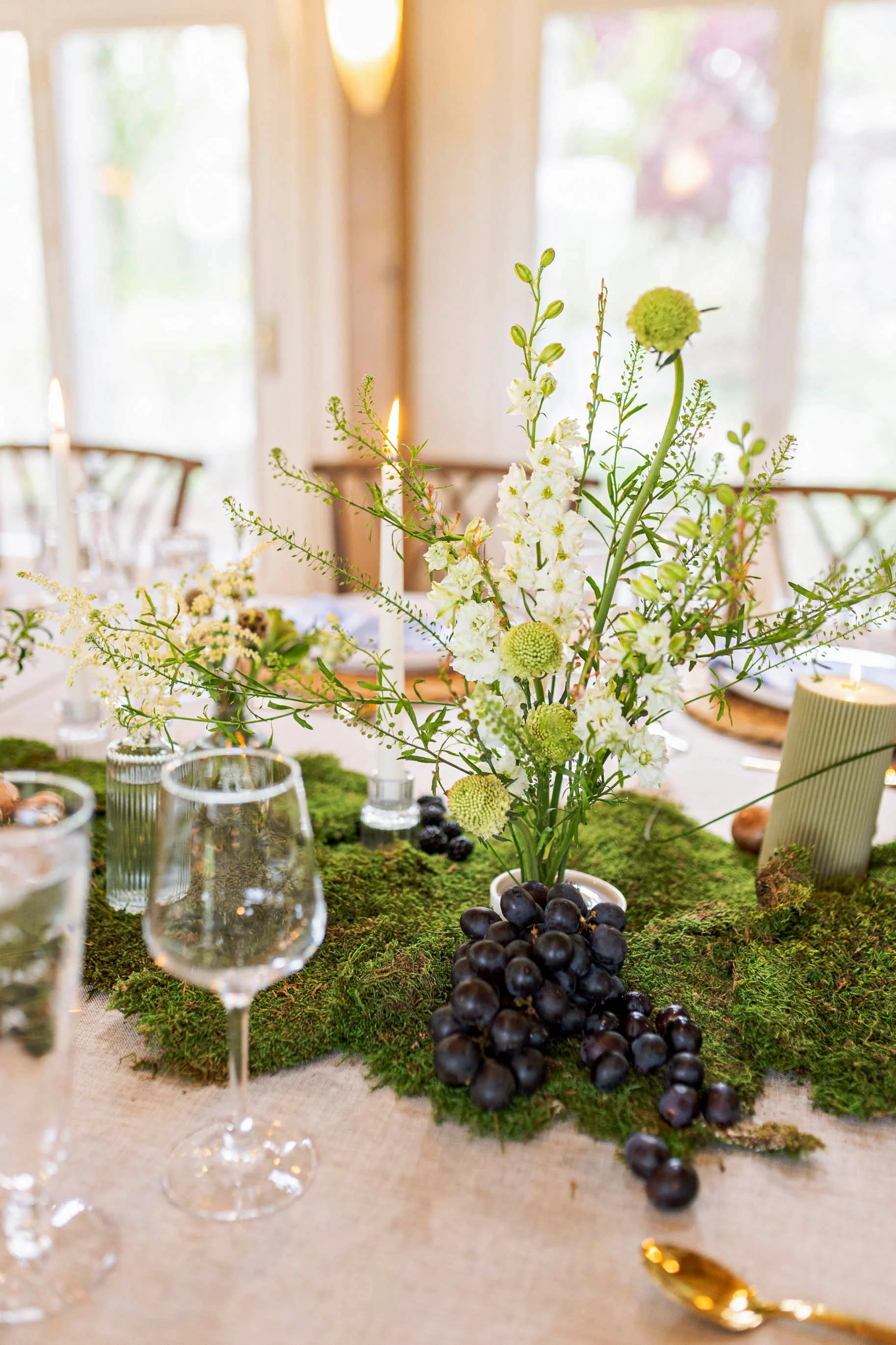 Table centerpiece with white and green flowers, black grapes, and candles on a mossy green table runner.