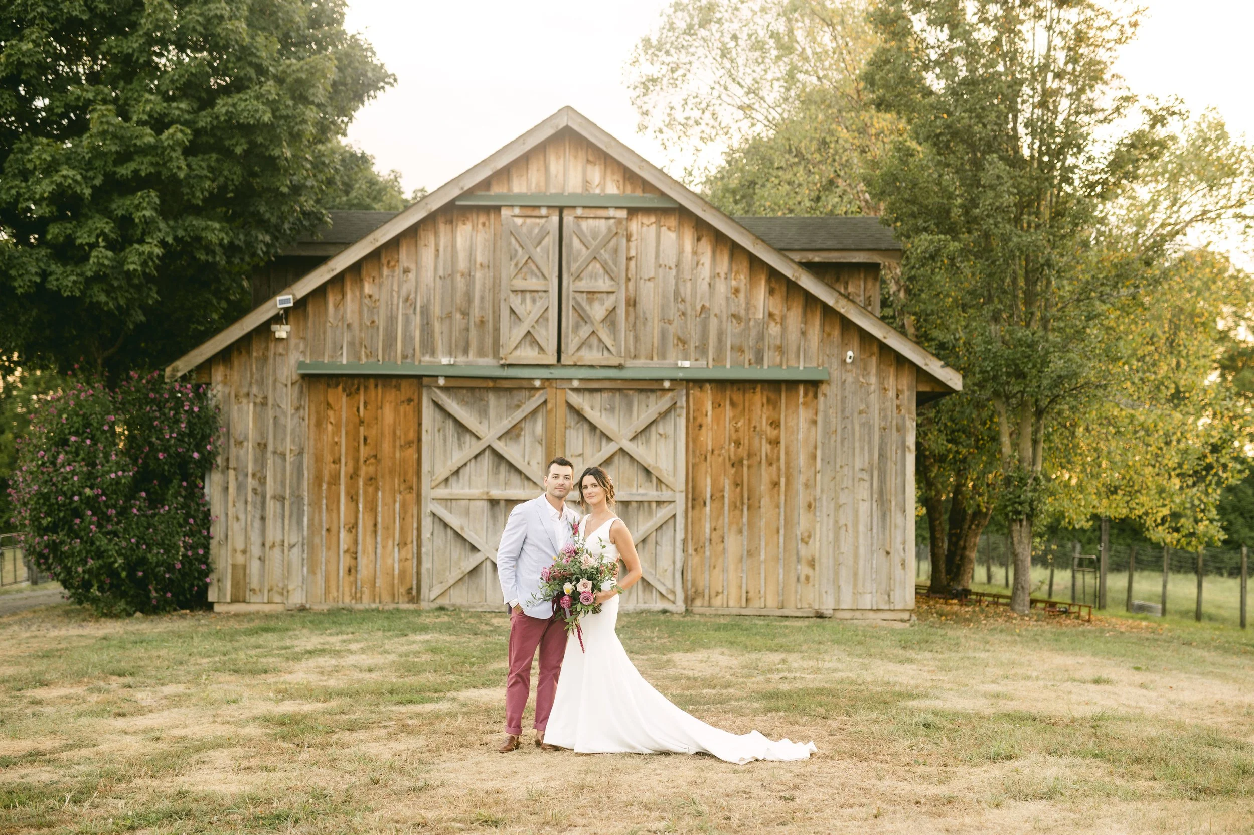 A bride and groom standing in front of a rustic wooden barn during a wedding photoshoot, with trees and a grassy field in the background.