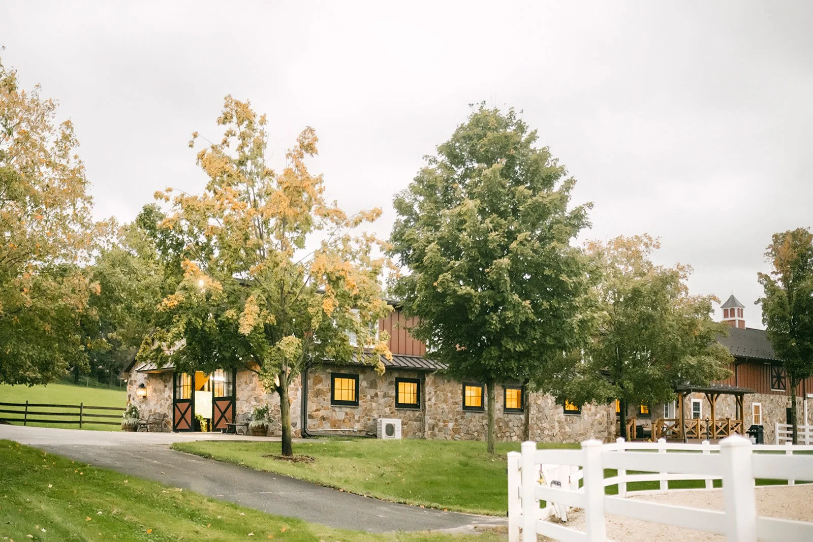 A rustic stone and wood barn with yellow-lit windows, surrounded by green grass and trees with autumn-colored leaves, and a white fence in the foreground.