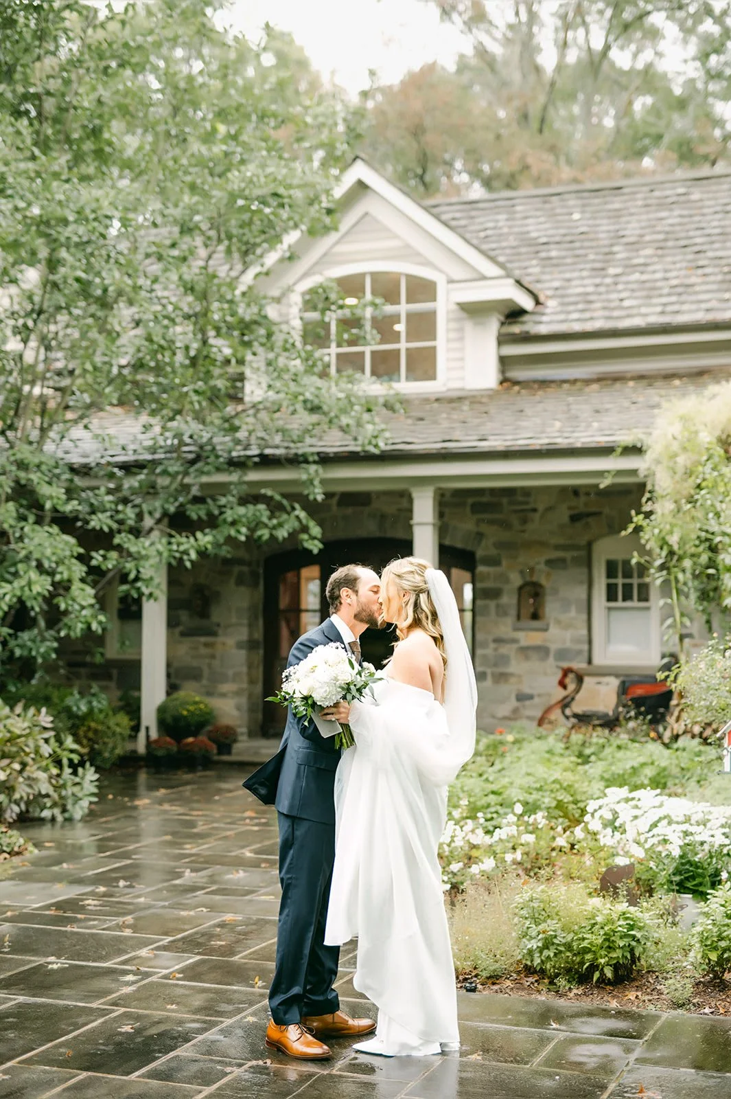 A newlywed couple sharing a kiss while holding a bouquet on a stone patio in front of a house surrounded by greenery and trees.