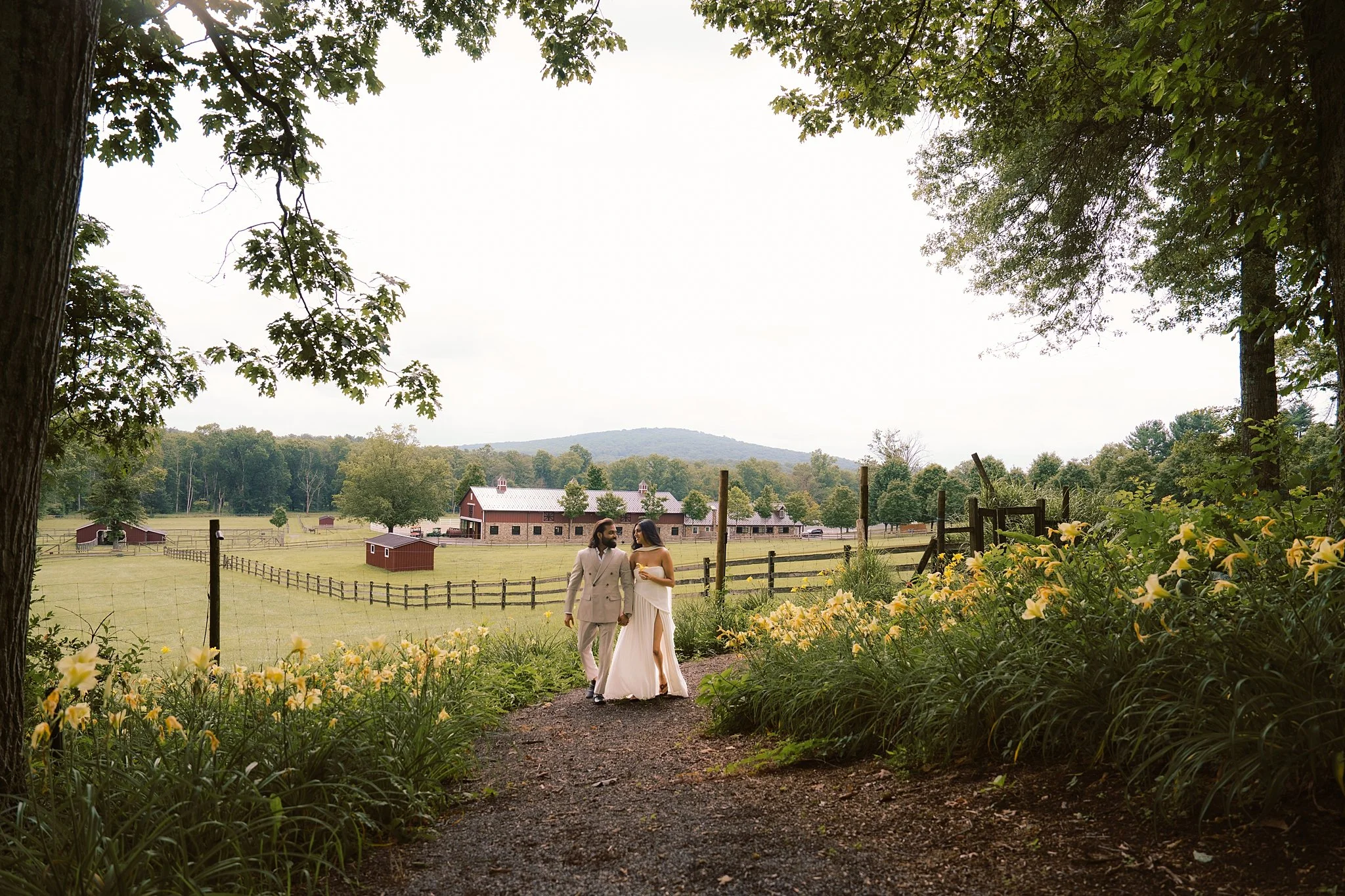 A couple in wedding attire walking hand in hand on a gravel path through a lush garden with yellow flowers, countryside farm buildings, and trees in the background.