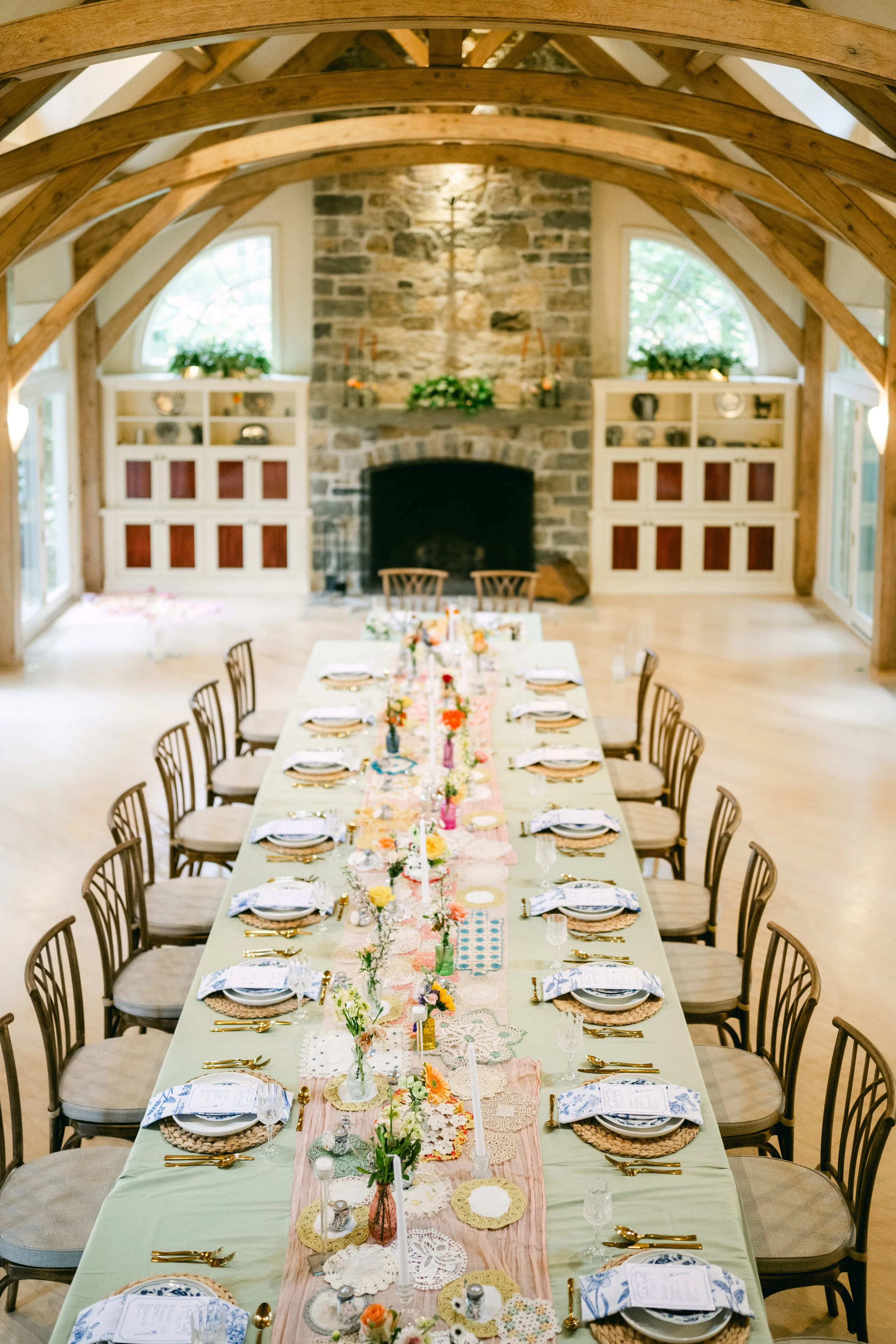Long dining table set for a celebration with floral centerpieces, candles, and place settings in a rustic wood ceiling room with a stone fireplace.