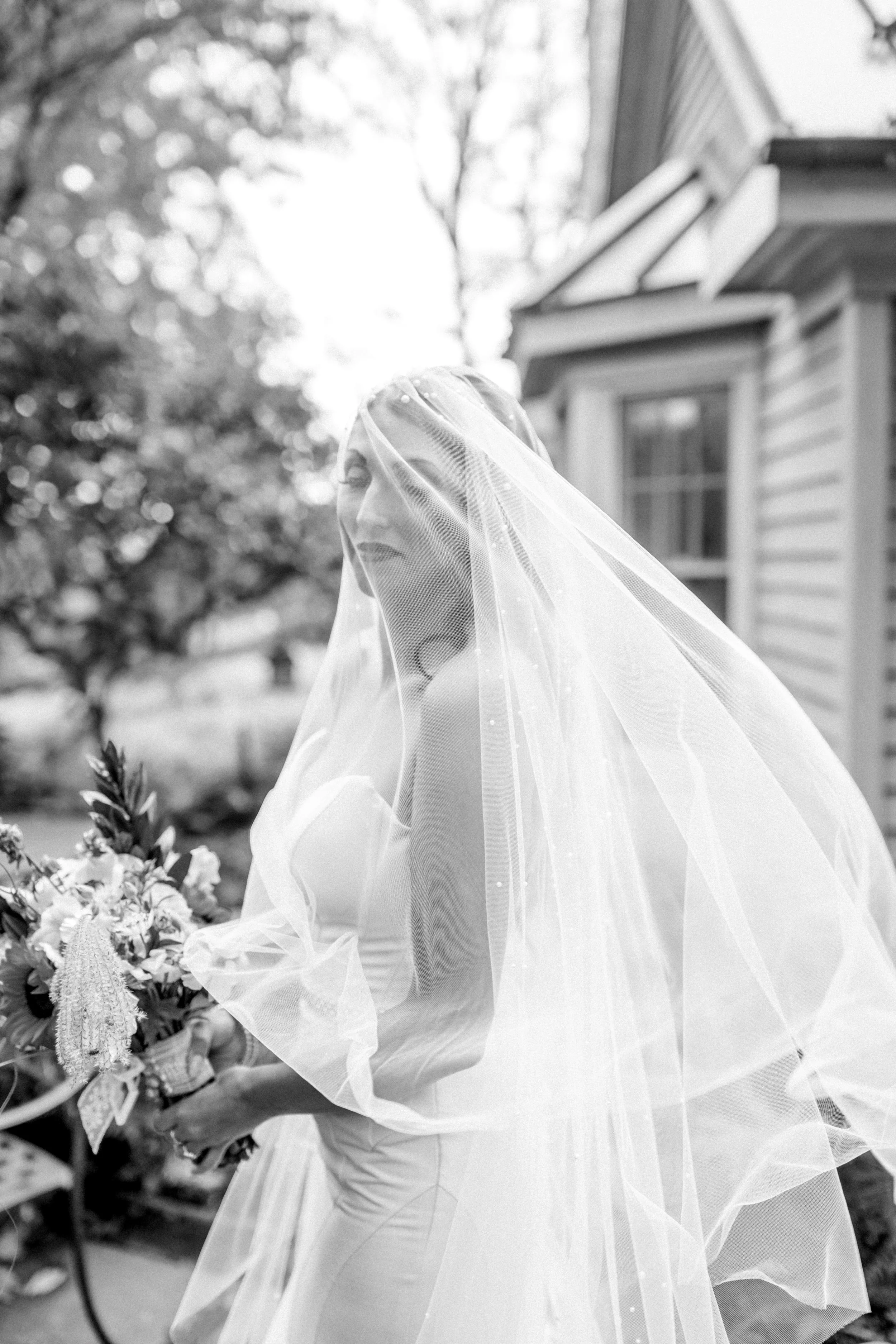 A woman with a veil over her face wearing a wedding dress, holding a bouquet of flowers outdoors near a house.