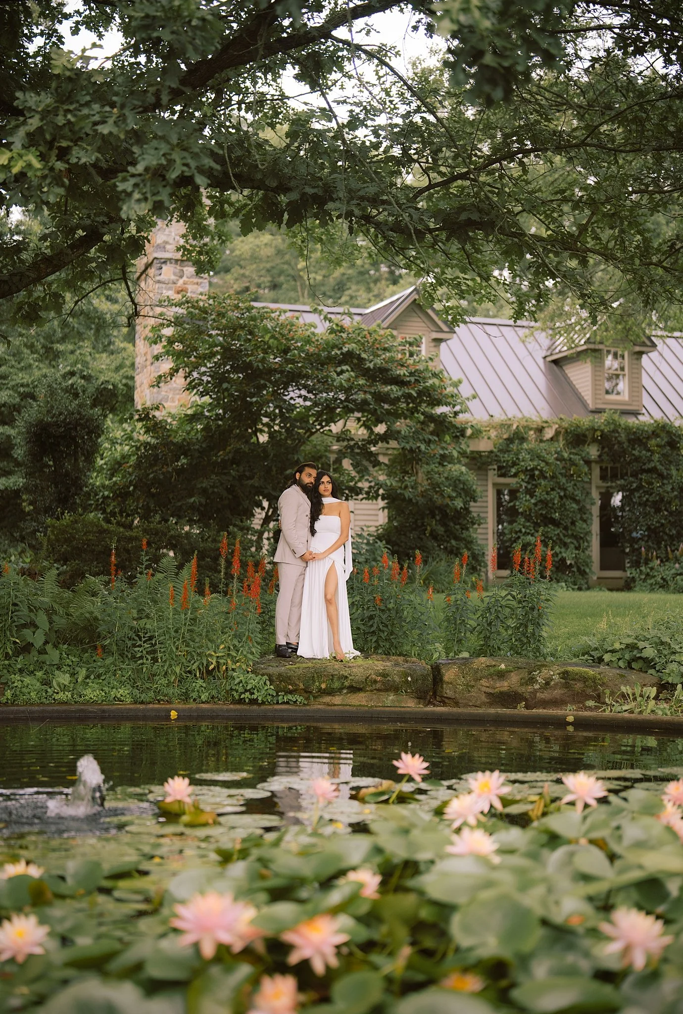 A bride and groom standing hand-in-hand by a pond with pink water lilies, surrounded by lush greenery and a house in the background.