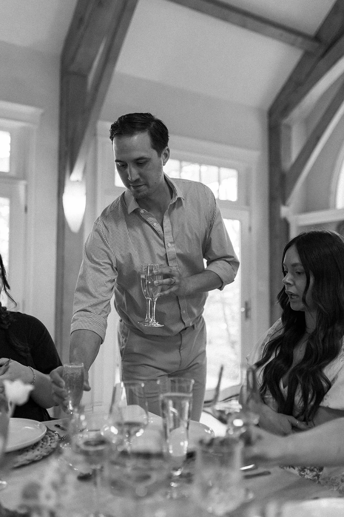 A man in a checkered shirt is pouring a drink at a table with women seated around it, indoors with large windows and wooden beams.