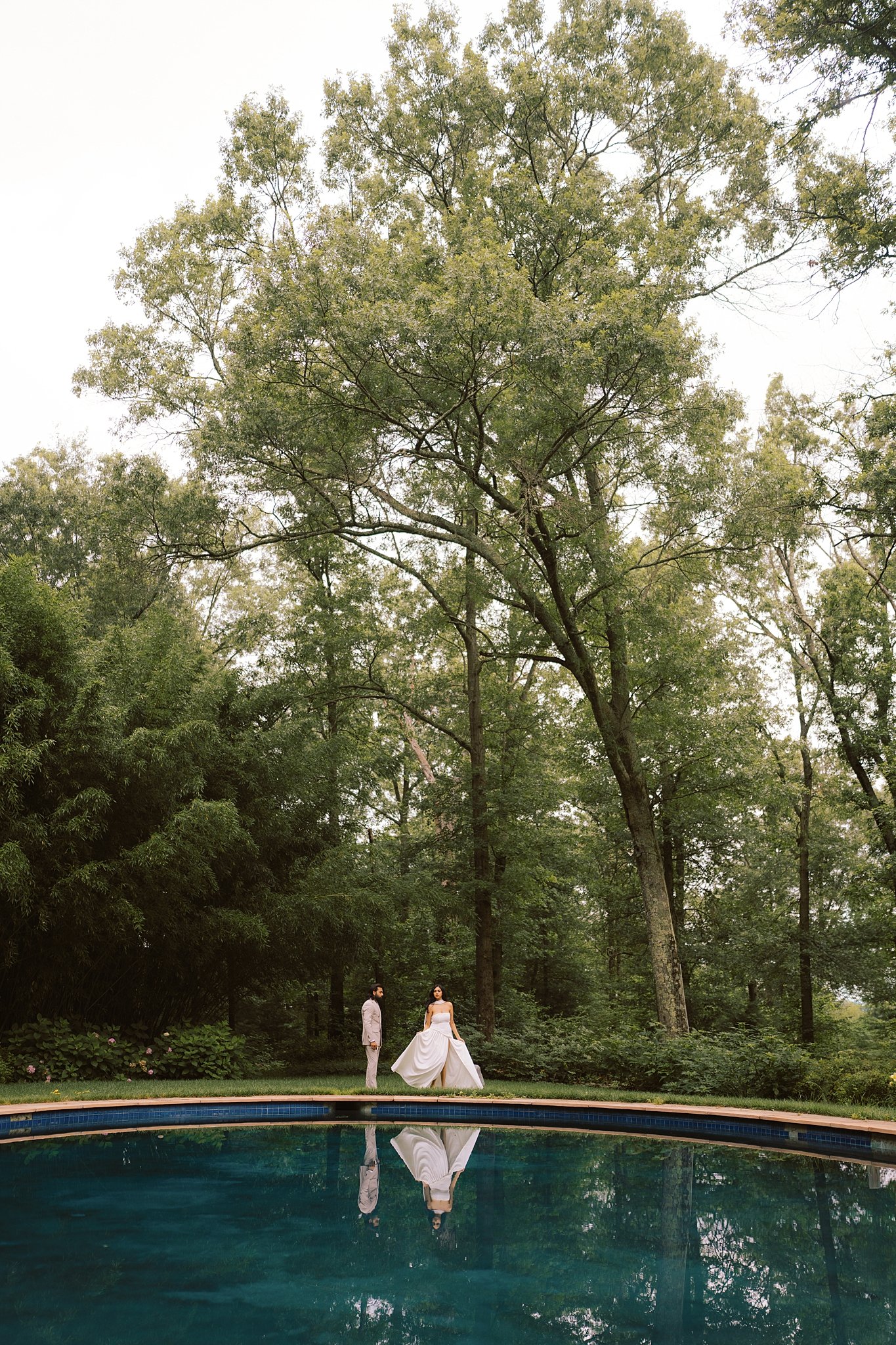 A bride and groom standing by a swimming pool surrounded by trees, with the bride in a white wedding gown and the groom in a light suit, their reflection visible in the pool water.