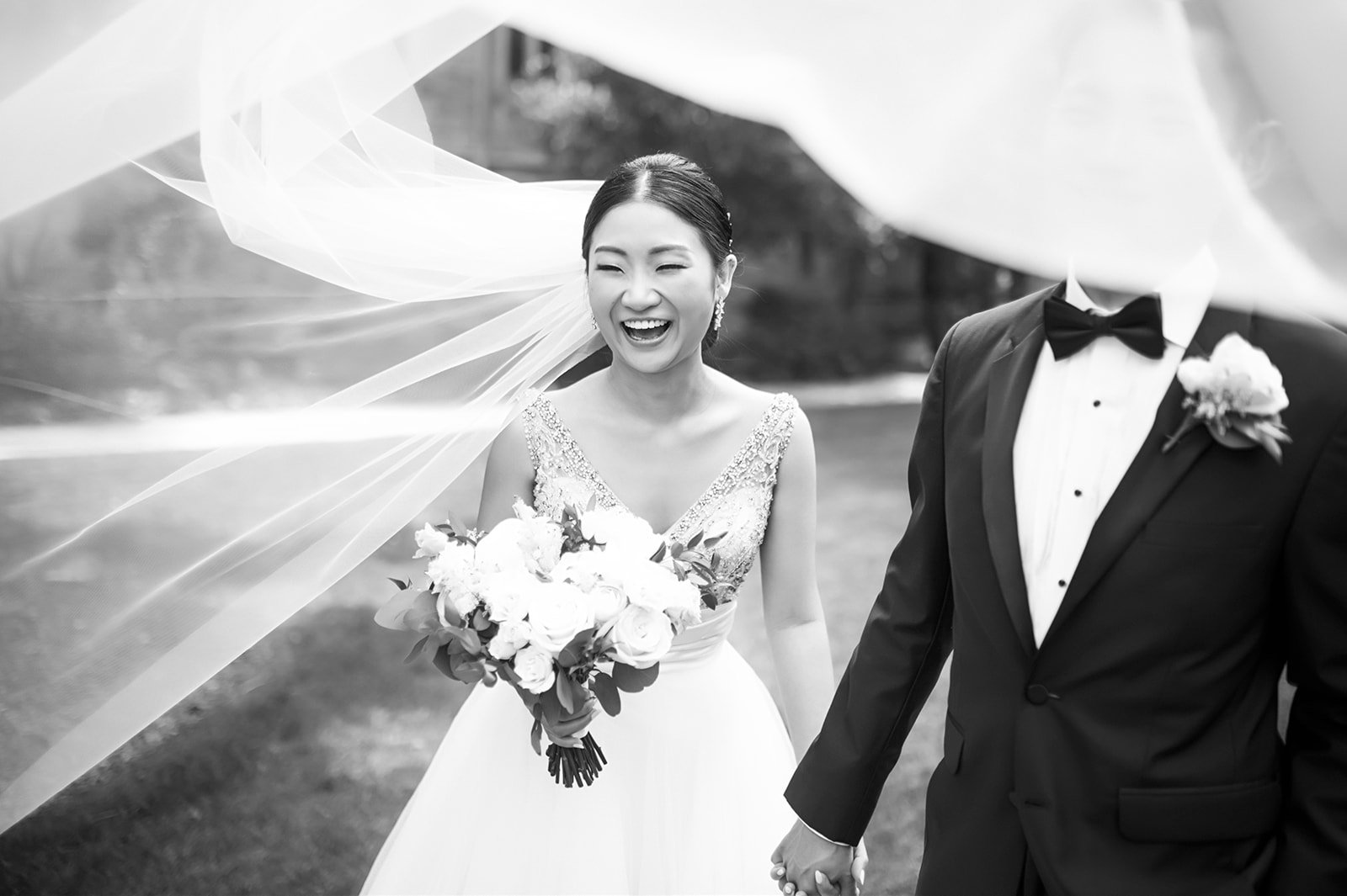 A bride in a wedding dress holding a bouquet of flowers, smiling, standing outdoors with a groom in a tuxedo, holding hands.