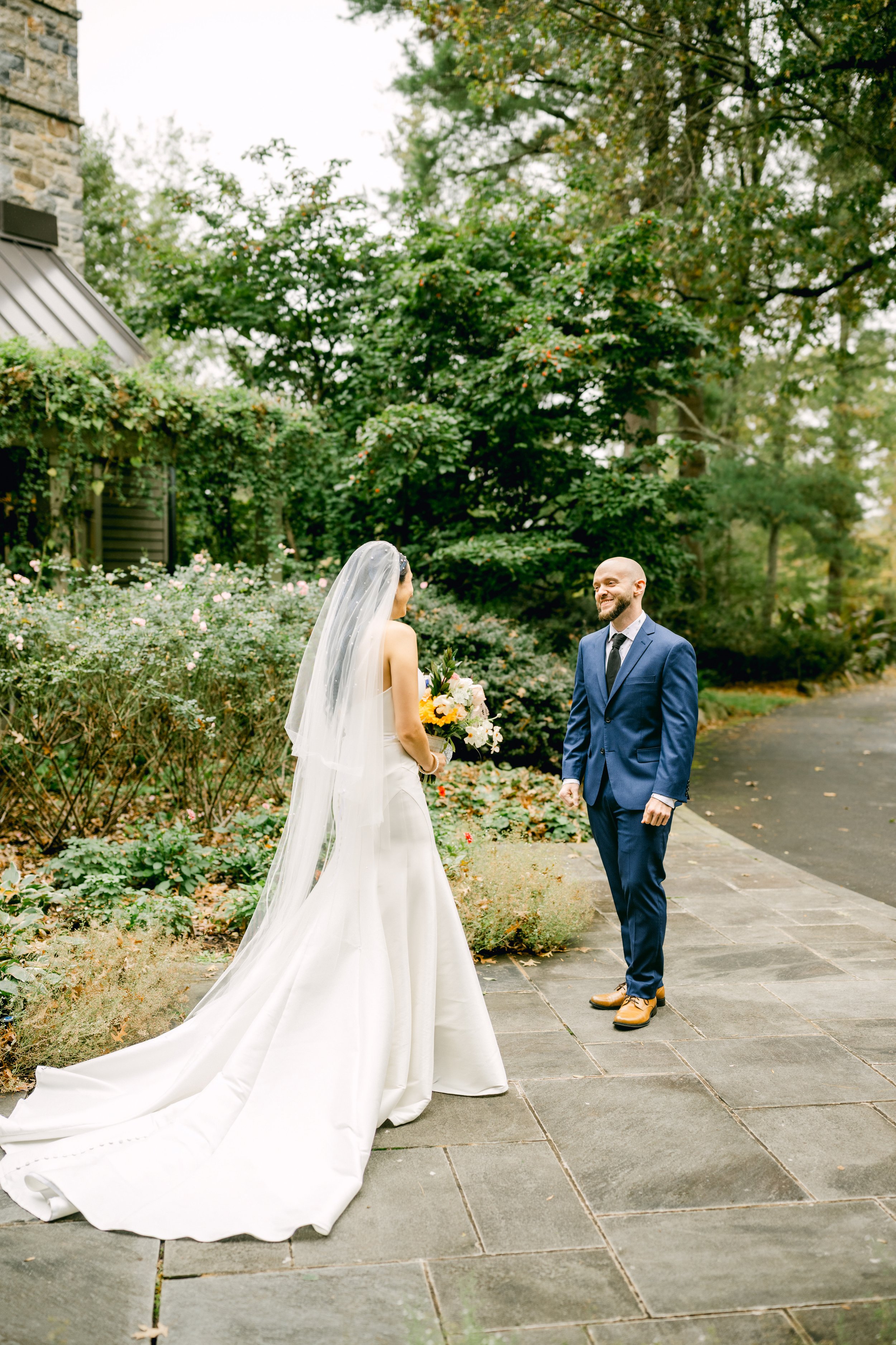 Bride in white wedding dress with veil holding a bouquet talking to groom in blue suit and brown shoes outdoors with trees and shrubs.
