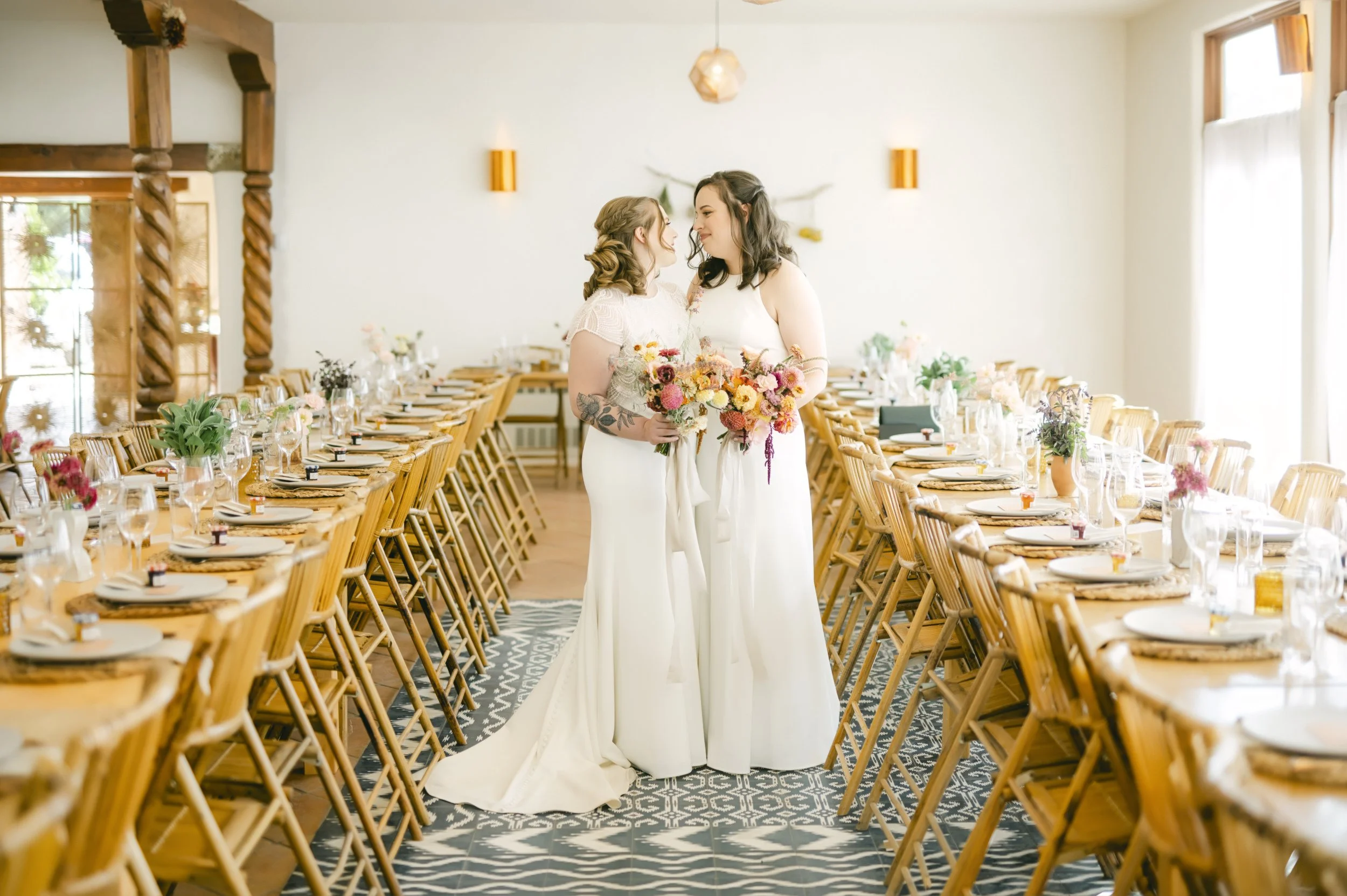 Two women in wedding dresses standing close together, holding bouquets of flowers, inside a decorated dining hall with wooden chairs and tables set for a celebration.
