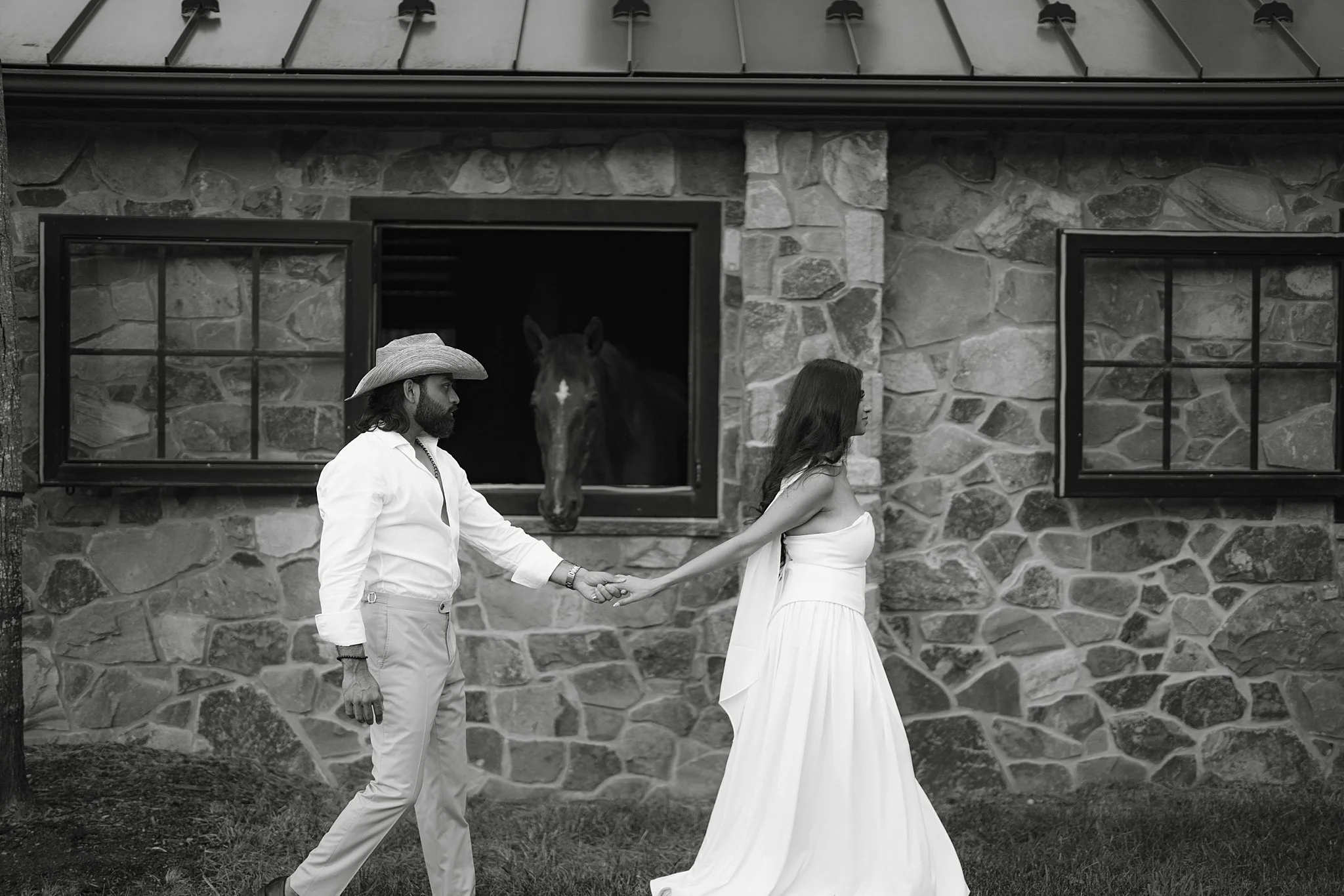 A black-and-white photo of a man and a woman holding hands outside a stone barn. The man wears a cowboy hat and white shirt, and the woman wears a strapless white dress. A horse looks out from the barn window between them.