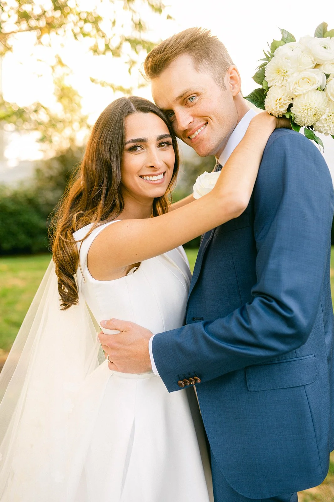 Happy bride and groom embracing outdoors during wedding, woman in white dress and man in blue suit, smiling, with flowers, trees, and sunlight in background.