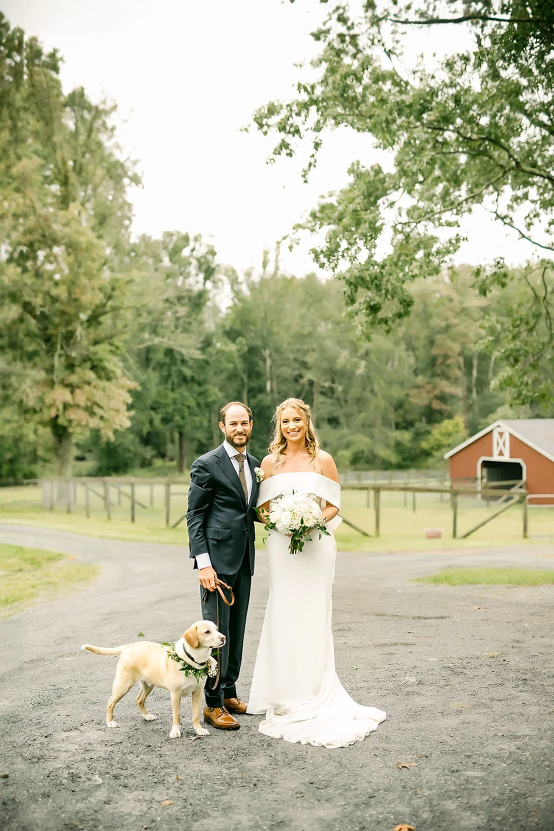 A wedding couple standing outdoors on a dirt path with a dog, surrounded by trees and a red barn in the background.