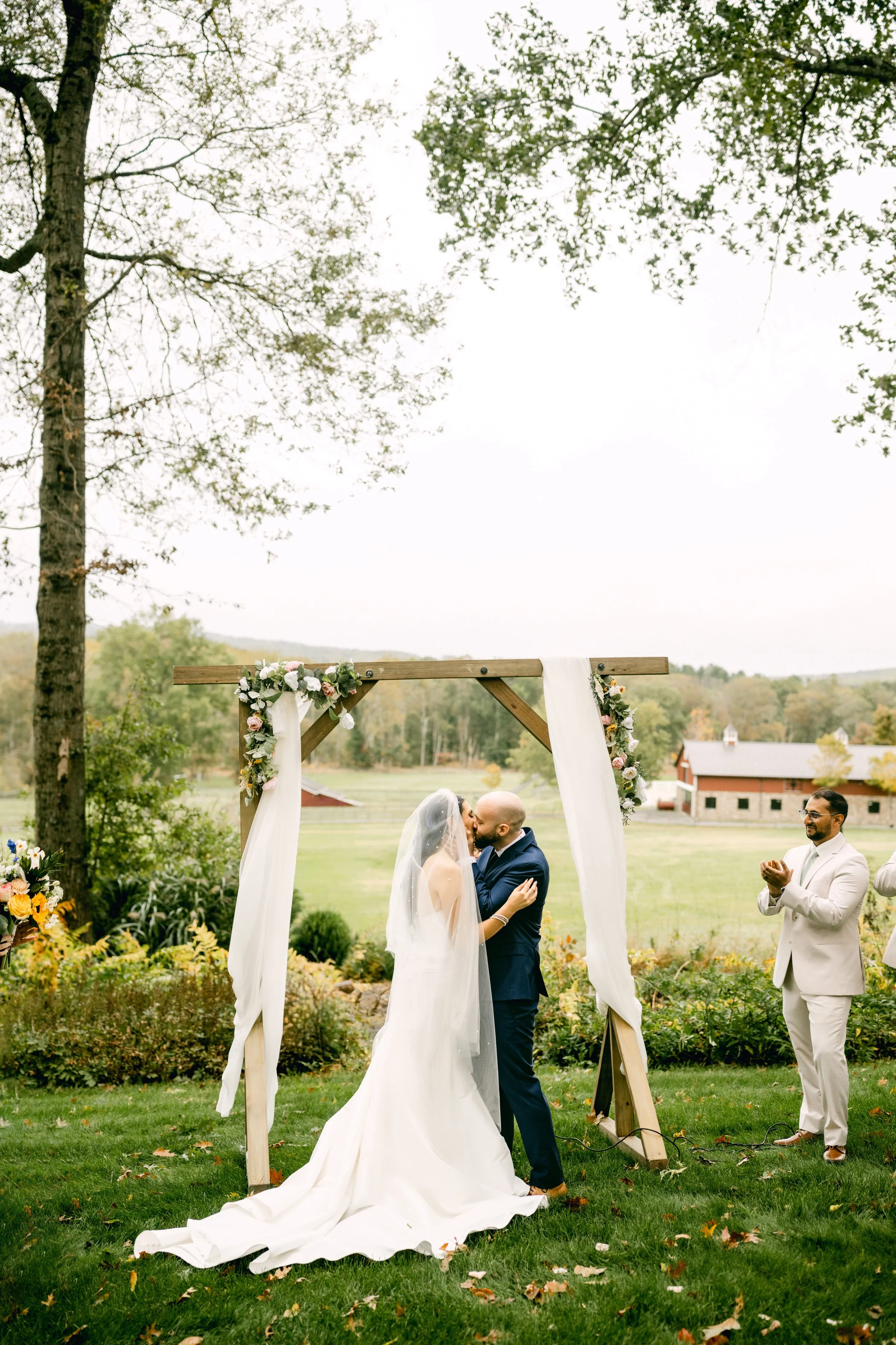 Couple getting married outdoors, sharing a kiss under a wooden arch decorated with flowers and white fabric, with a person clapping nearby, in a lush green setting with trees and buildings in the background.
