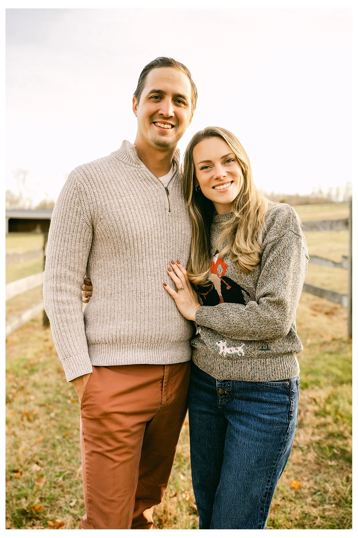 A young couple standing outdoors on a grassy area, smiling and embracing each other. The man has short dark hair and is wearing a beige sweater and brown pants. The woman has long light brown hair, is wearing a patterned sweater and blue jeans, and has her hand resting on the man's chest.